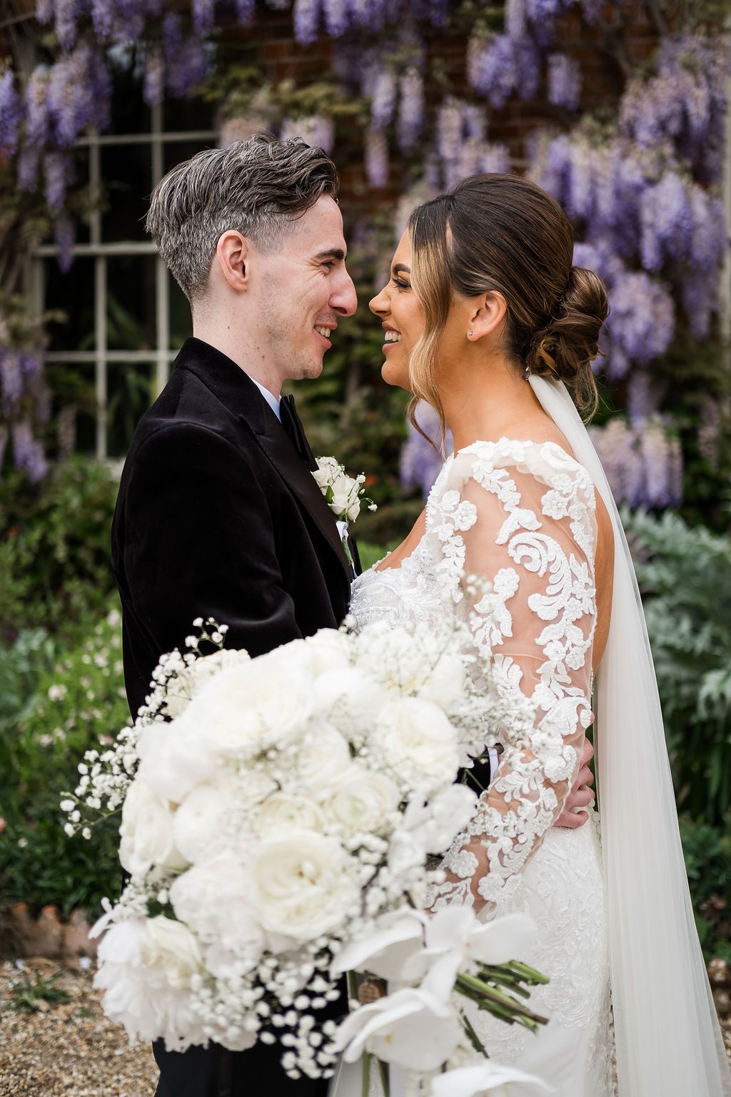 Newlywed couple embraces in garden setting with wisteria blooms and white floral bouquet.
