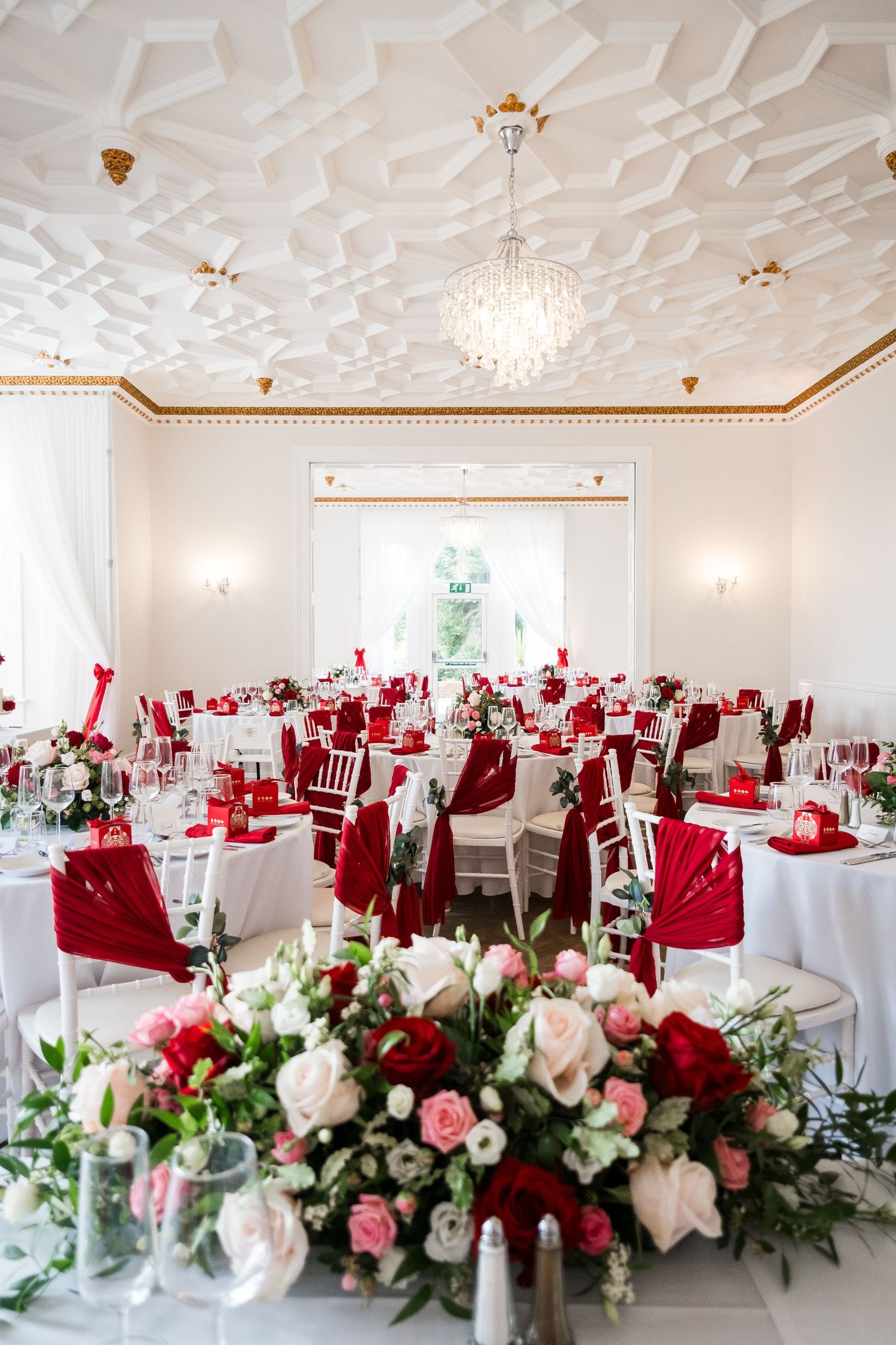 Elegant wedding reception venue with white walls, crystal chandeliers, and red and white floral centerpieces on round tables.