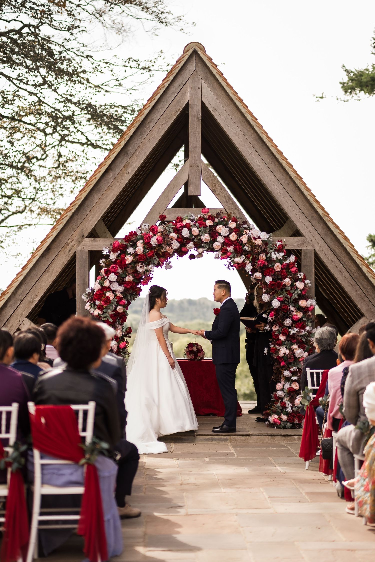 Wedding ceremony under a wooden A-frame pavilion decorated with red and pink floral arch.