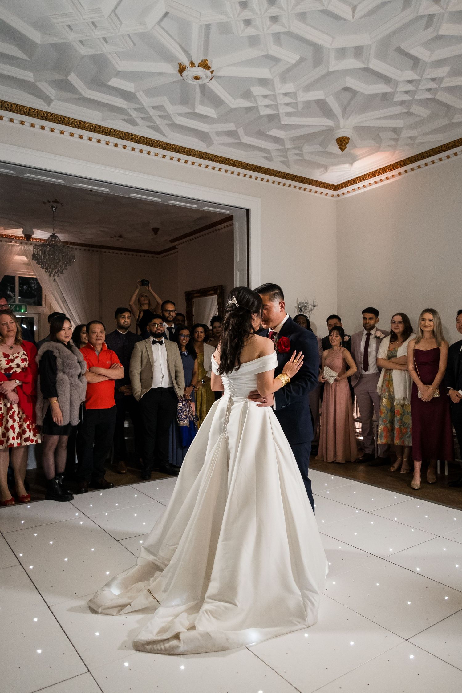 A couple shares their first dance on an illuminated white dance floor while wedding guests watch from the sides.