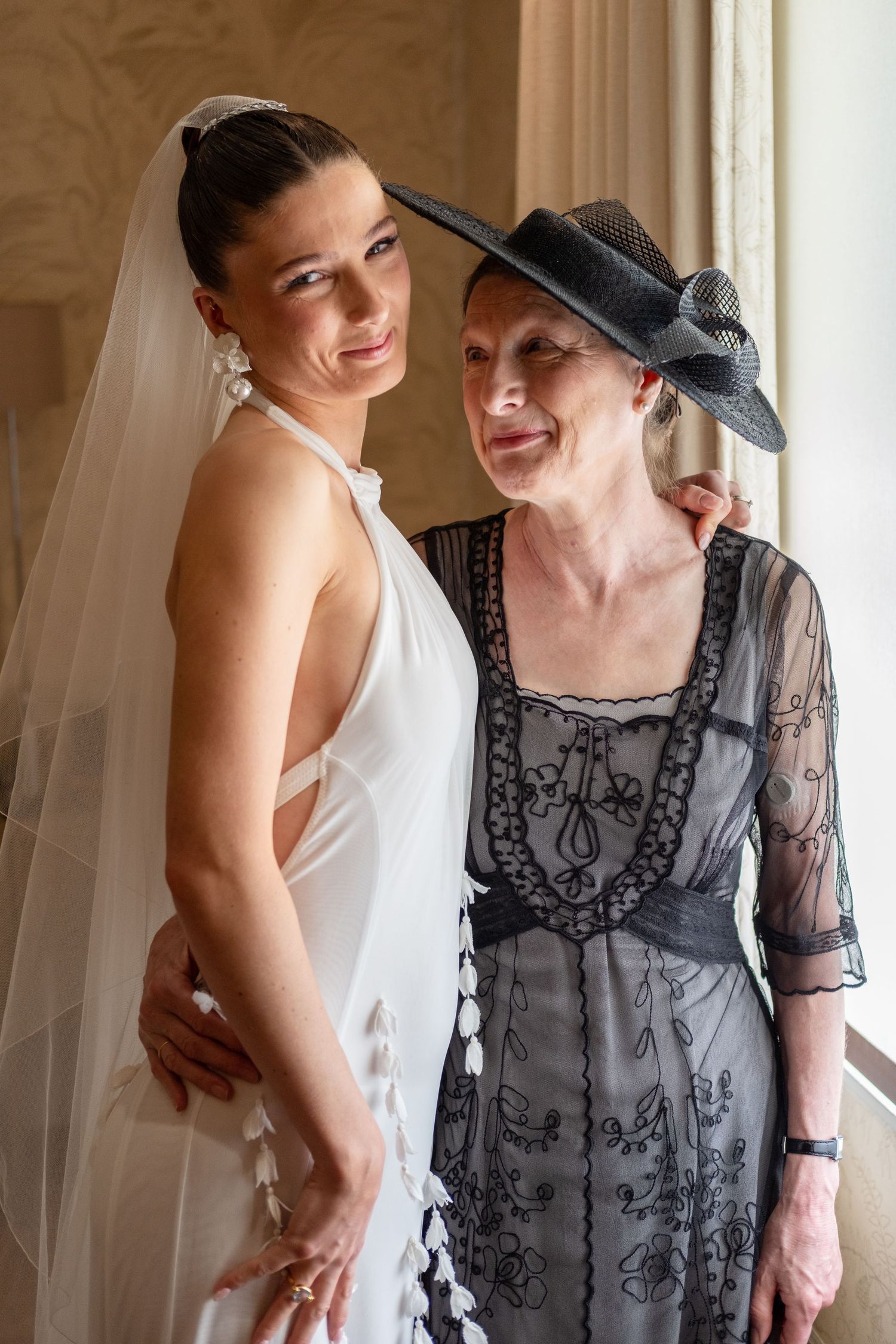 Bride in white gown and veil poses with older woman in black lace dress and hat at elegant wedding venue.