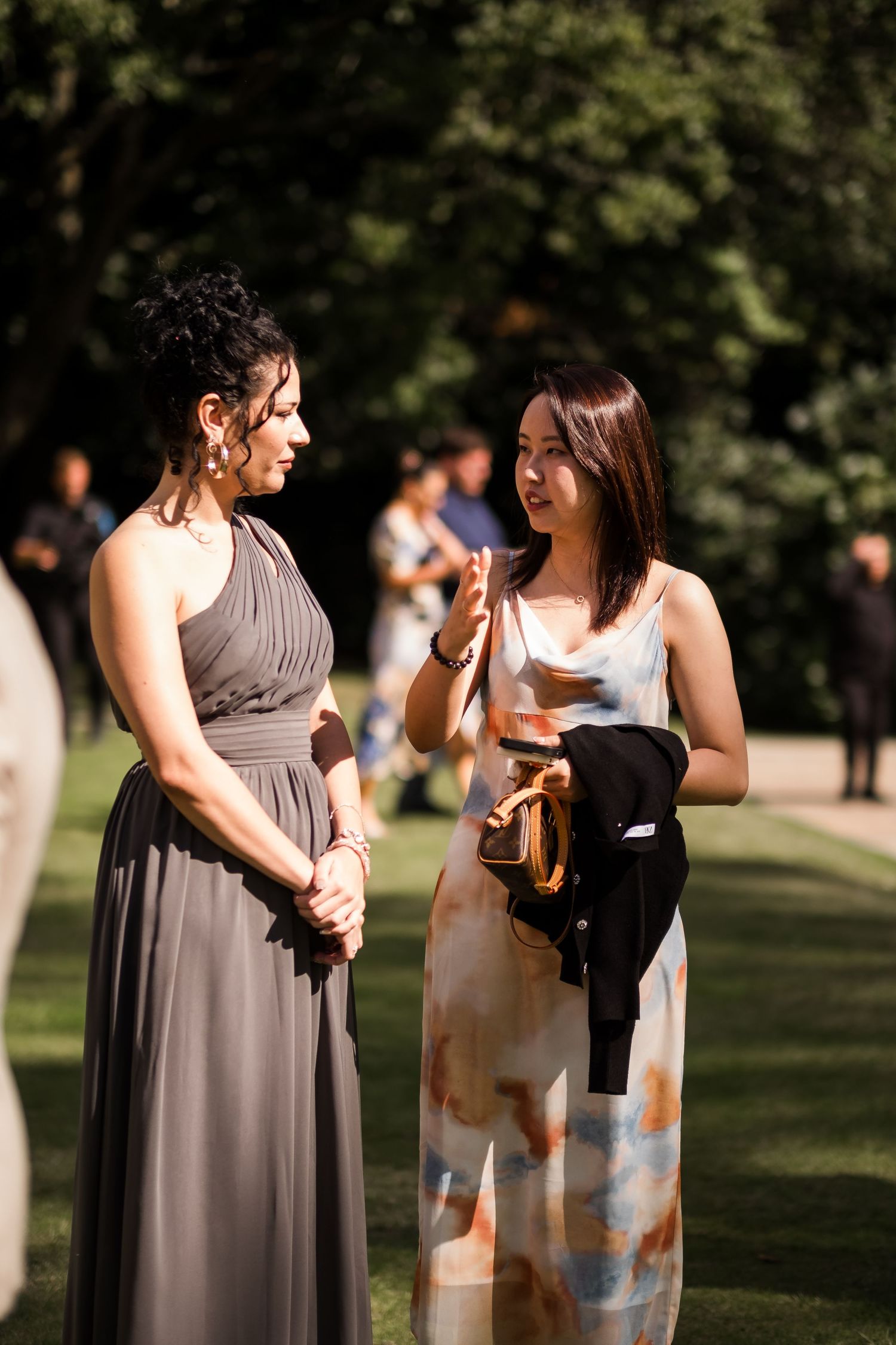 Two people in elegant dresses share a candid moment during an outdoor event in a sunlit park setting.