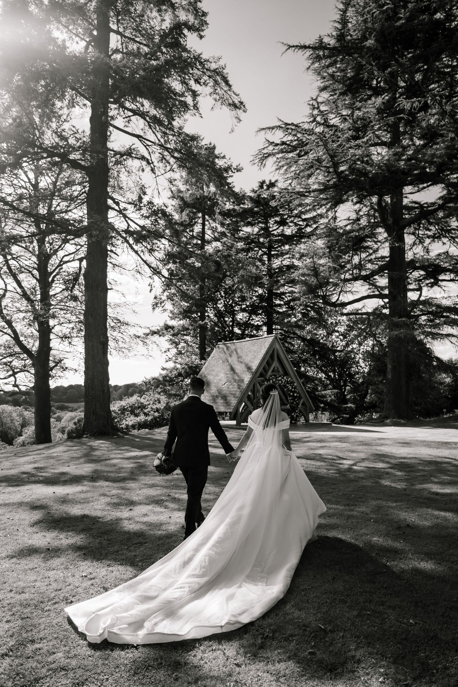 Black and white photo of bride and groom walking hand in hand toward a chapel surrounded by tall trees.