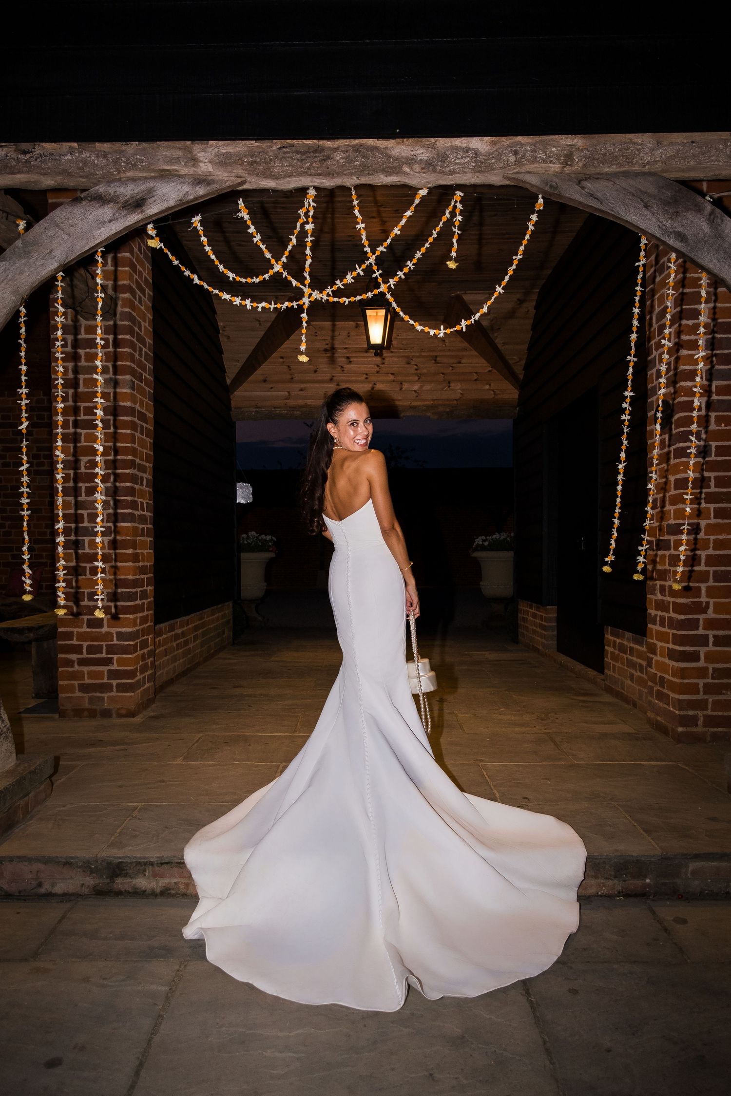 Bride in elegant white backless mermaid gown with train poses under fairy lights at rustic brick venue at night.