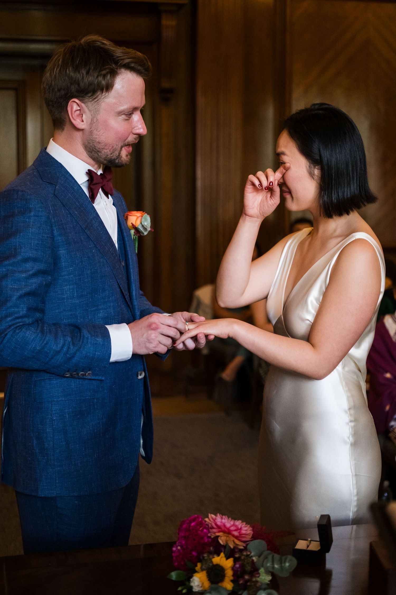 Emotional bride wiping tears while groom places wedding ring on her finger during intimate ceremony.