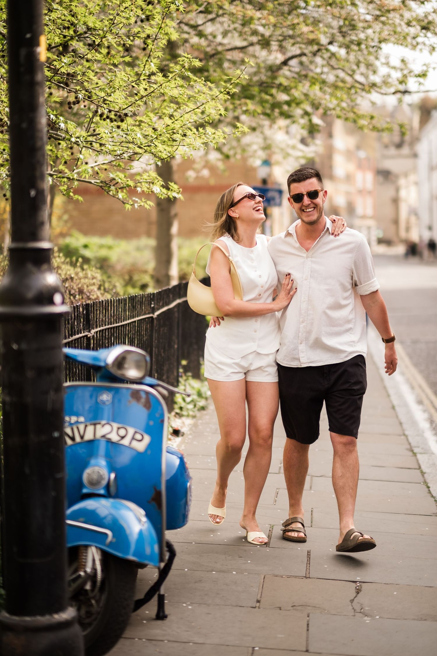 Happy couple walking arm in arm on a sunny cobblestone street beside a vintage blue Vespa scooter.