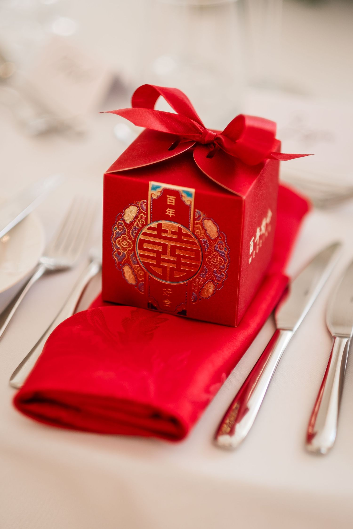 Traditional red Chinese wedding favor box with double happiness symbol and bow sits atop red napkin with silverware.