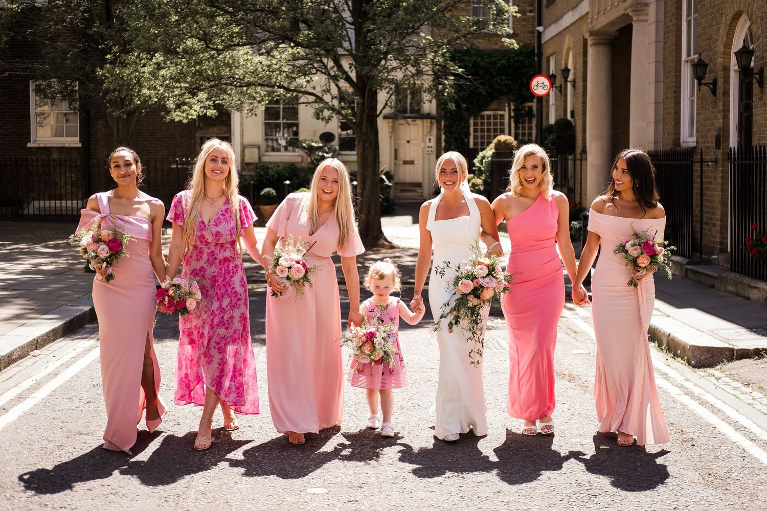 Bride and bridesmaids in pink dresses holding bouquets pose on a sunny street for a wedding photo.