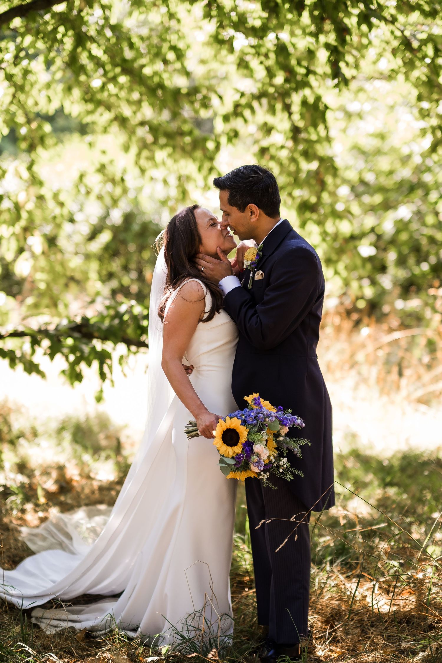 A romantic wedding couple shares intimate kisses in a sunlit wooded setting with vibrant sunflowers in their bouquet.