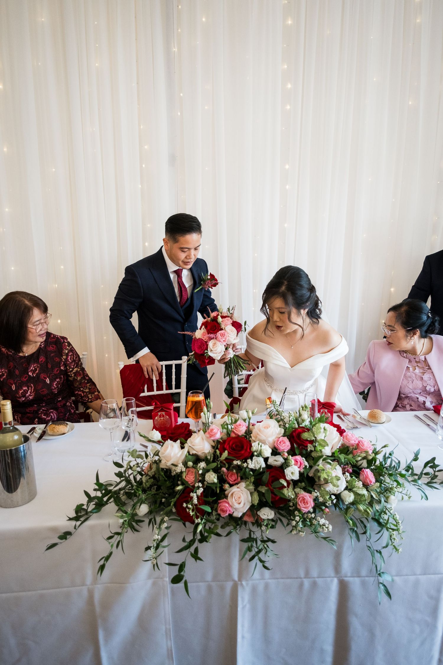 Wedding party members sign documents at a table decorated with pink and red floral arrangements during a ceremony.