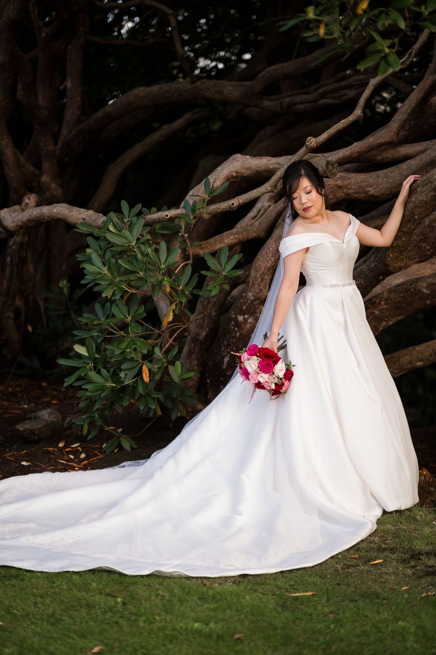 A bride in a flowing white wedding dress sits gracefully against a tree trunk while holding a vibrant pink bouquet.