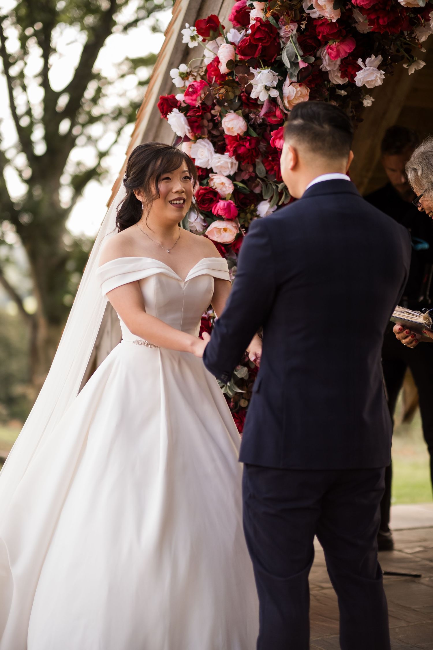 A couple exchanges vows under a floral arch decorated with red and white flowers during an outdoor wedding ceremony.