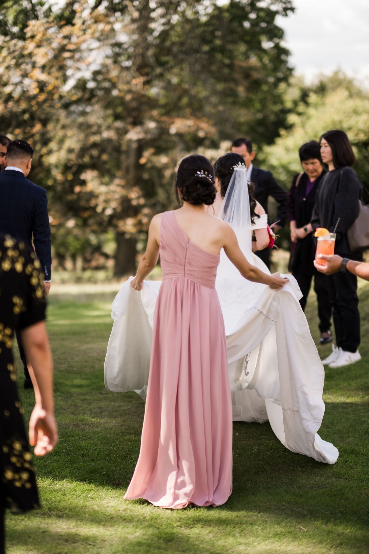 Wedding party members in pink bridesmaid dresses help adjust a white wedding gown train on a sunny lawn.