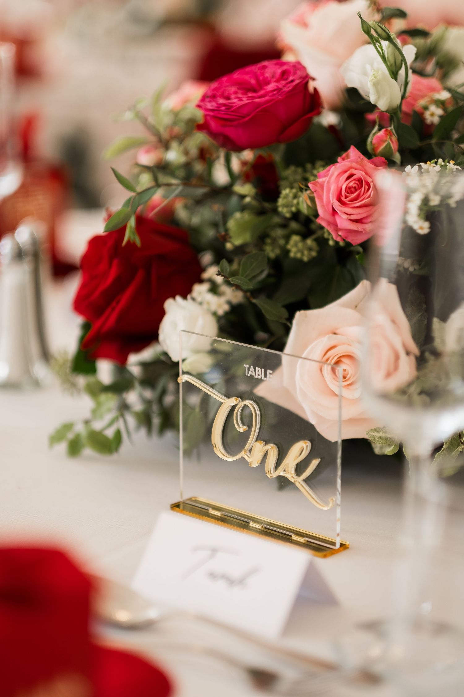 Elegant wedding reception table settings with pink and red roses, gold table numbers, and place cards in romantic lighting.