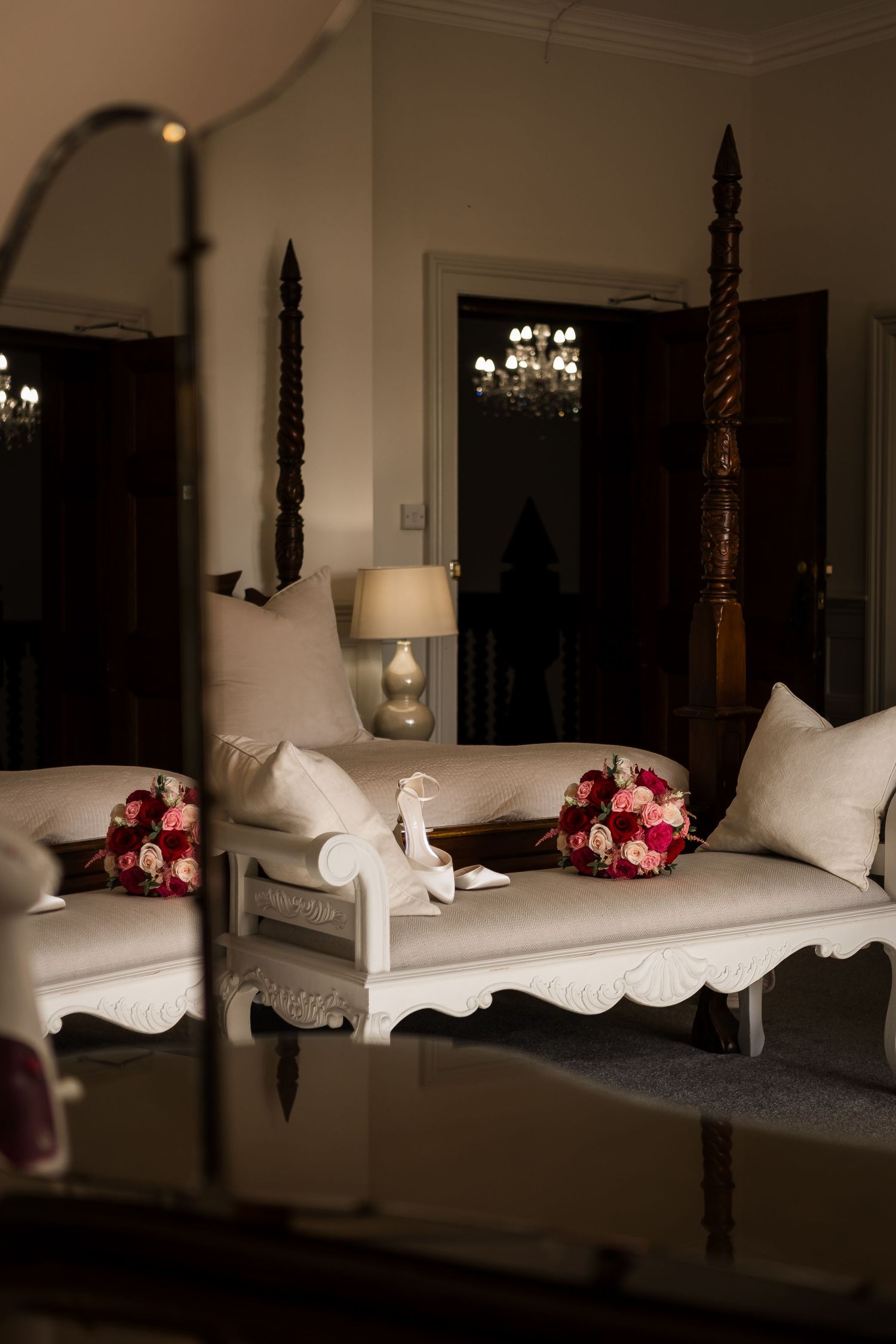 A luxurious four-poster bed with white linens and pink roses is illuminated by soft lighting in a romantic bedroom setting.