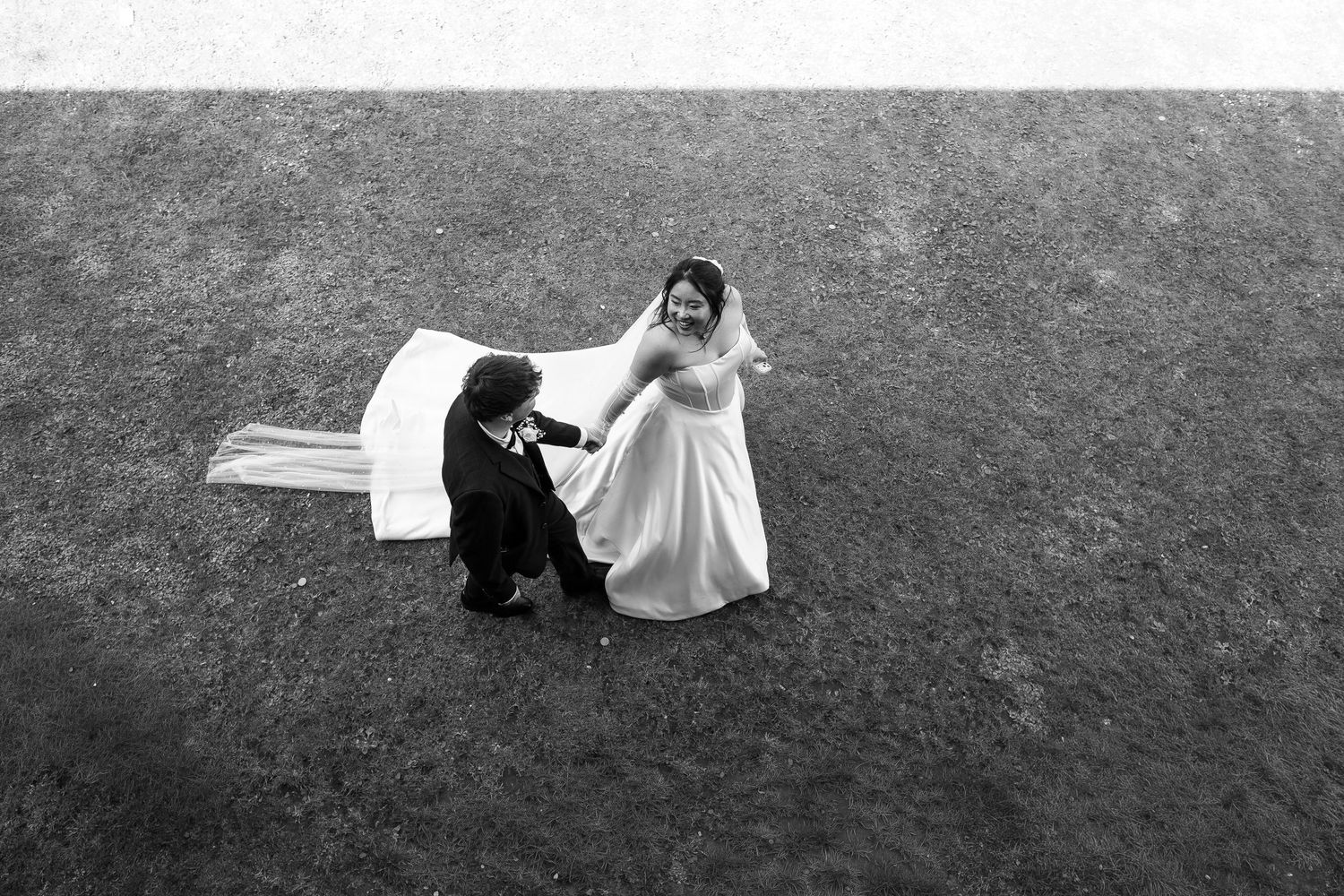 Aerial black and white photo of bride in white gown and groom in black suit dancing on green grass.