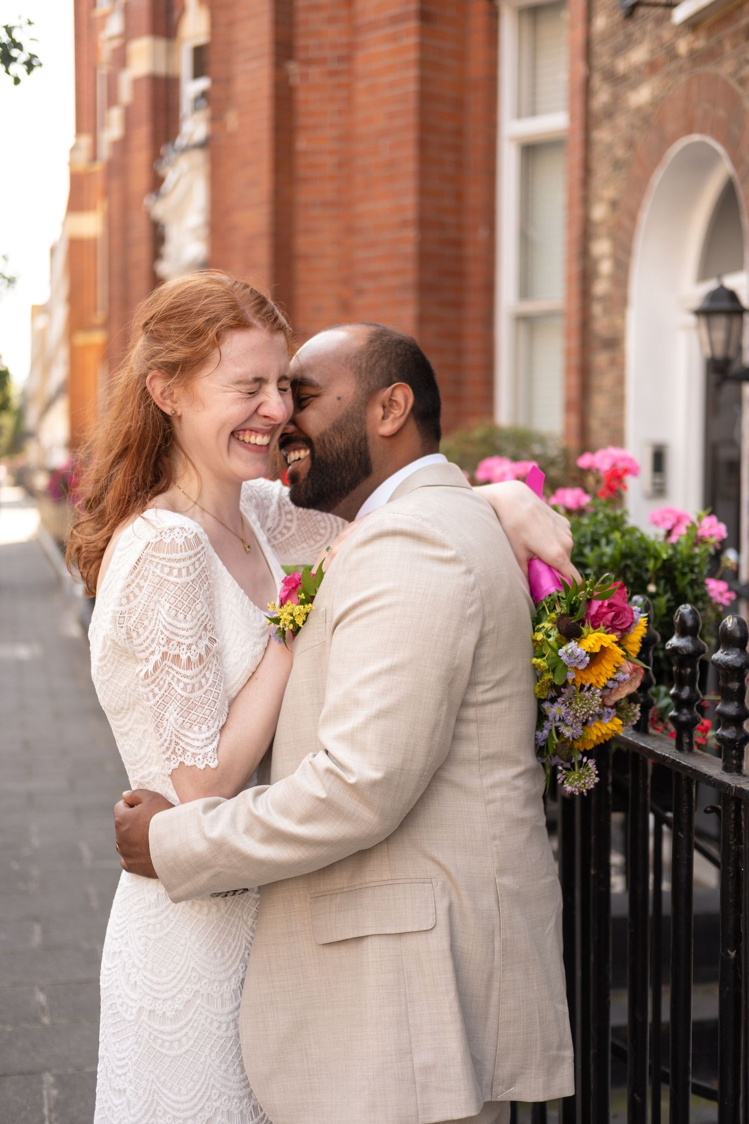 A joyful bride and groom embrace on a London street, she in white lace, he in a beige suit with a floral boutonniere.