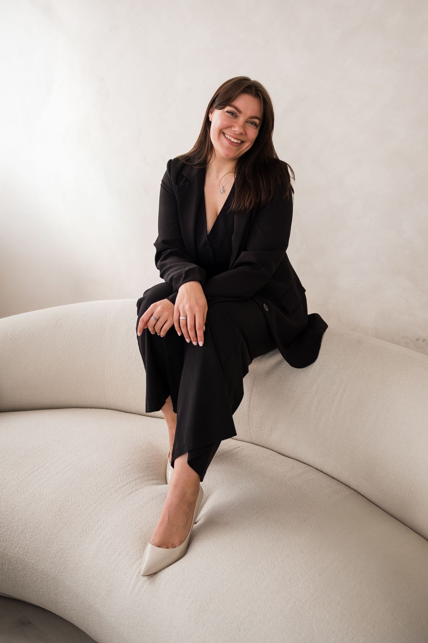 Smiling woman in black blazer and trousers sitting on a cream curved sofa in a bright minimalist room.