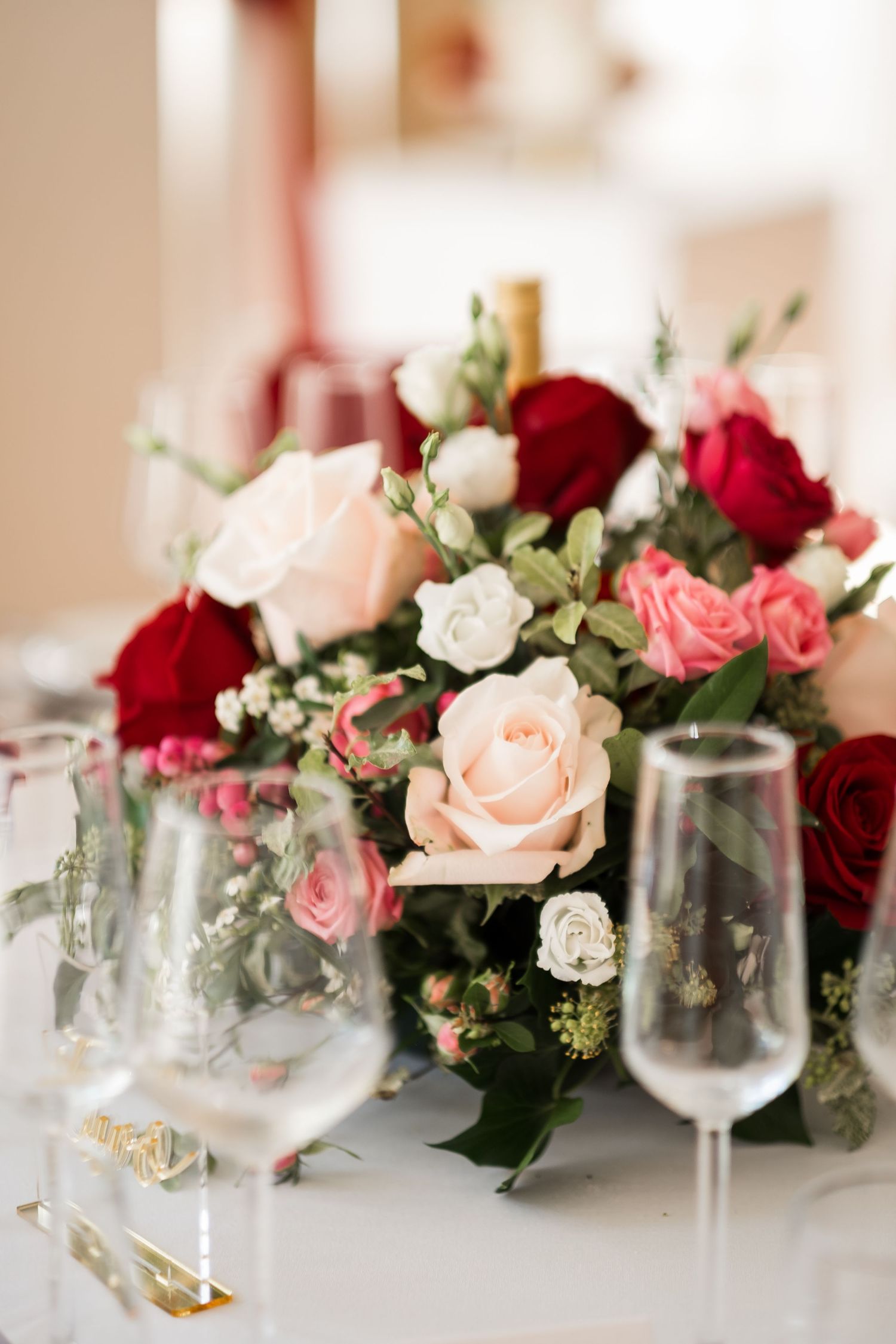 Elegant floral centerpiece with pink and red roses arranged with wine glasses on white tablecloth.