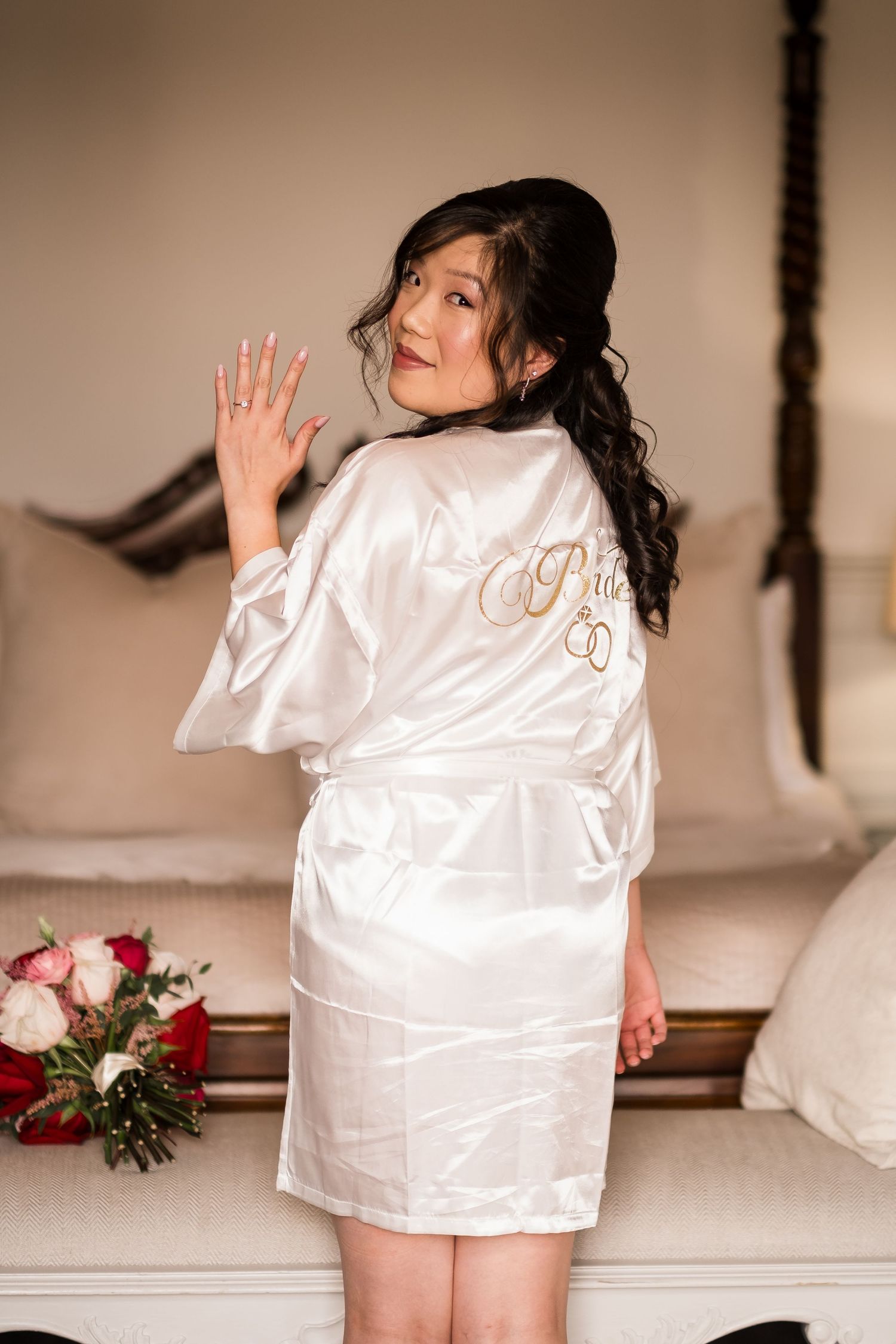 A person in a white silk robe poses with a bouquet of red and pink roses beside a couch.