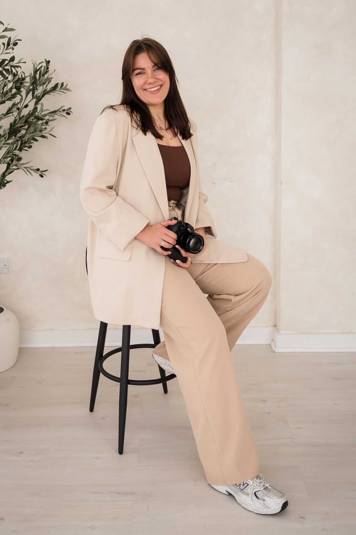 A photographer in beige business attire sits casually on black stools against a white studio backdrop.