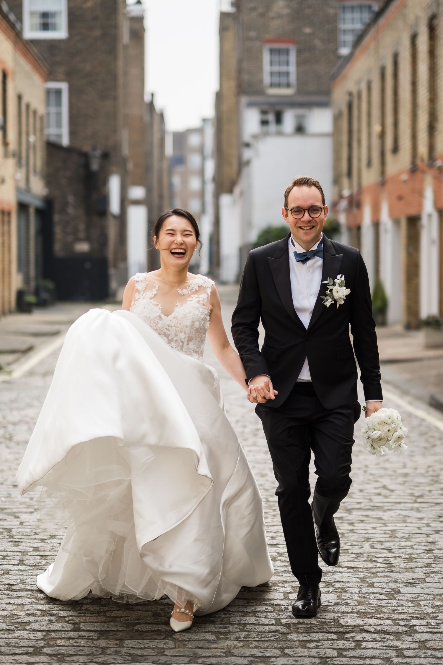 Joyful bride and groom walk hand-in-hand on cobblestone street, bride in white gown, groom in black tuxedo with bow tie.