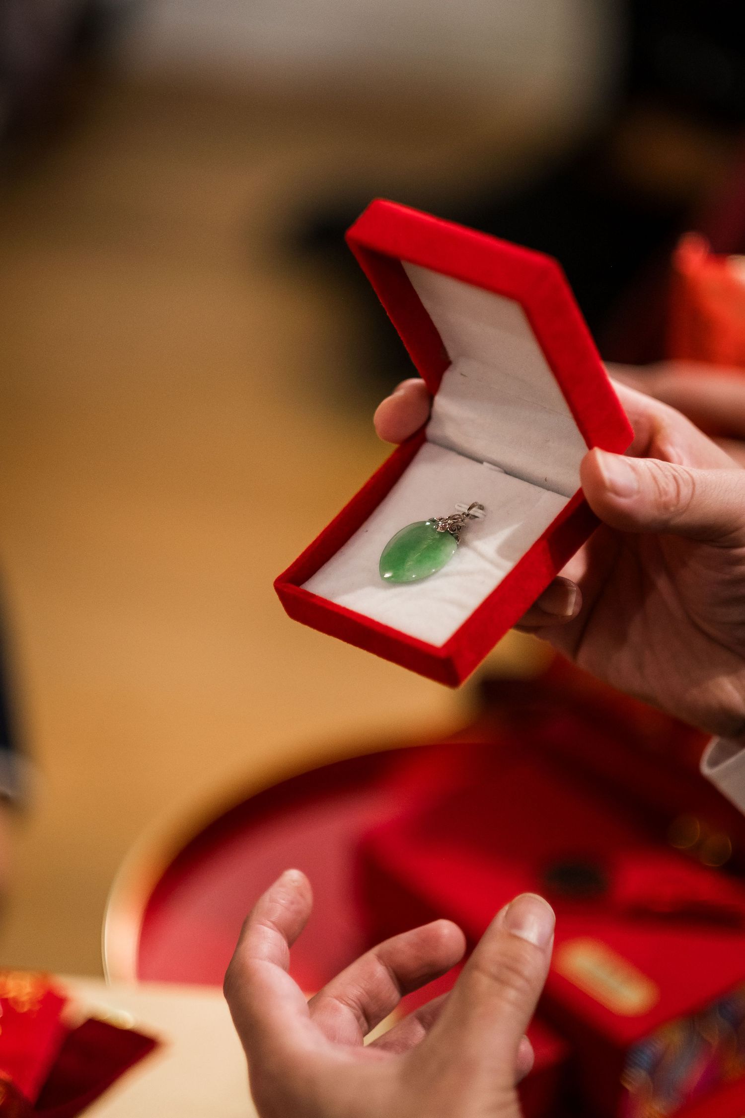 Hands opening a red jewelry box containing a green jade pendant against a warm-toned background.