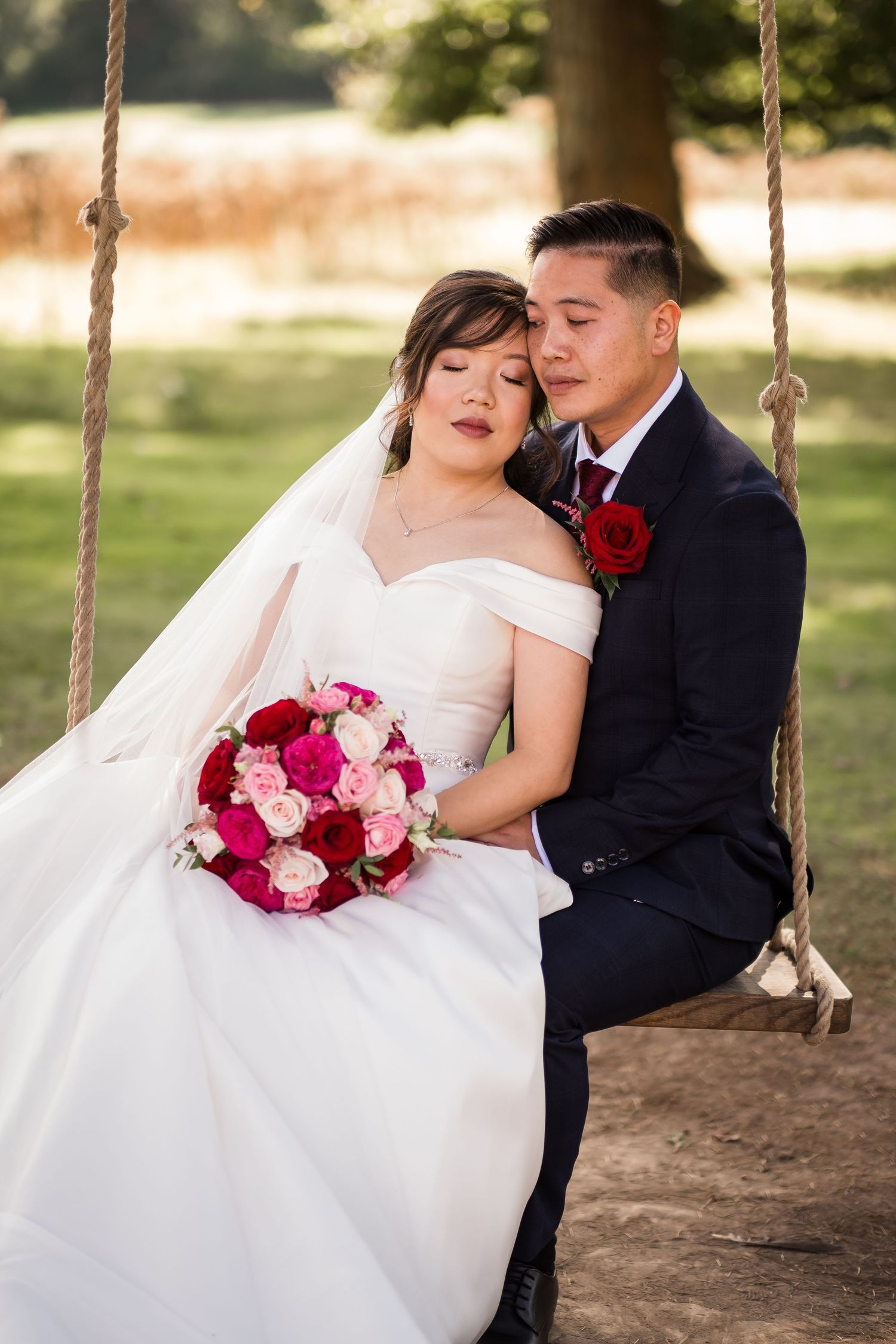A romantic wedding couple poses on a wooden swing with red and pink rose bouquets in an outdoor garden setting.