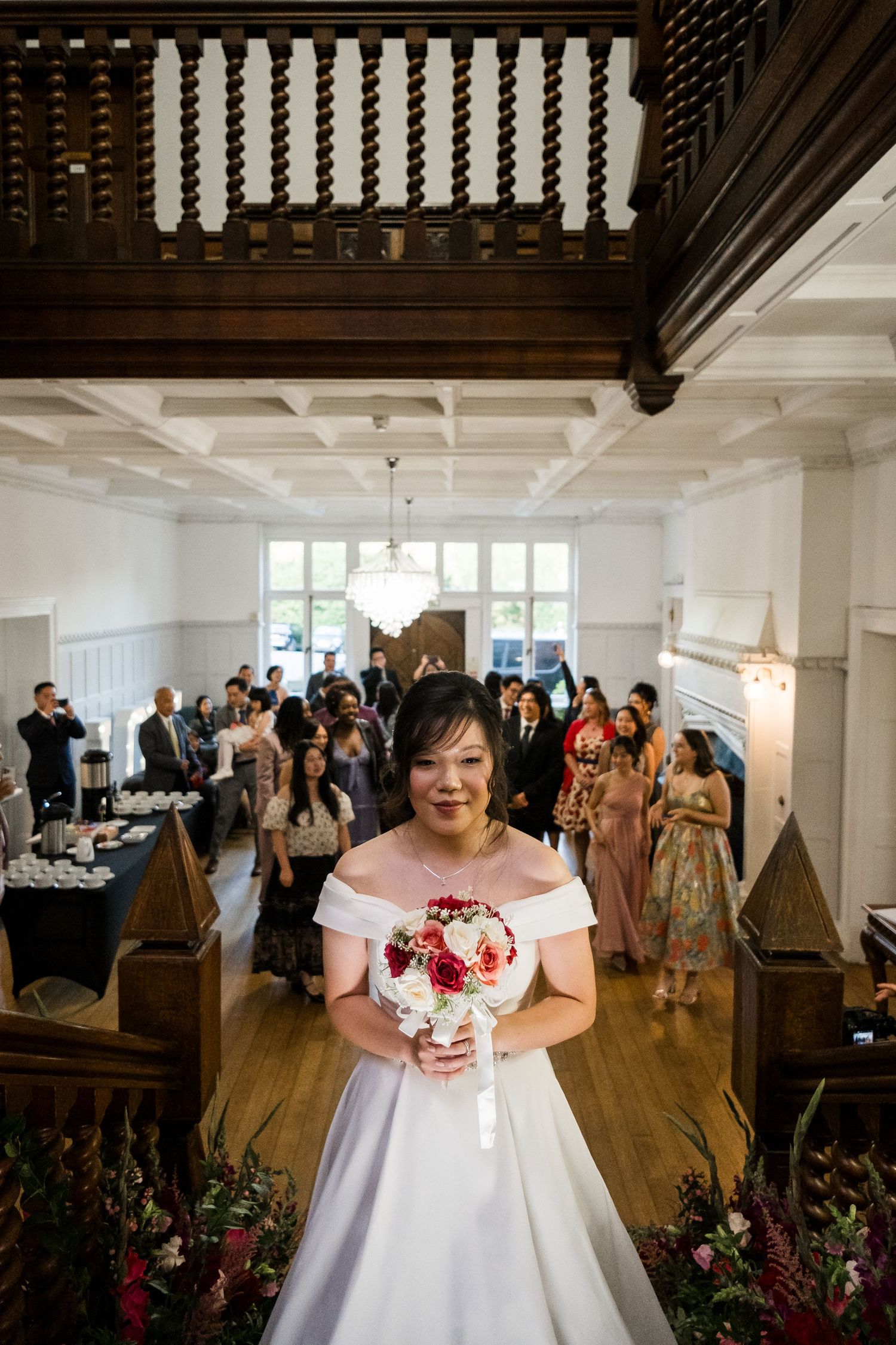 Wedding guests gather in an elegant white reception hall with wood beam ceilings and natural light.