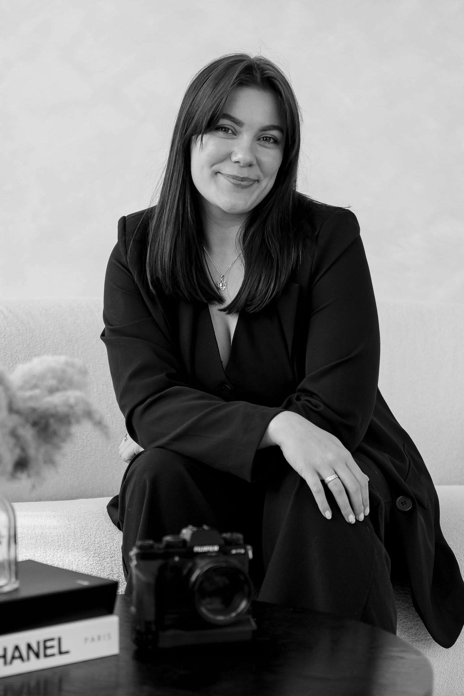 Professional black and white portrait of a person in business attire sitting with a camera and Chanel book on table.