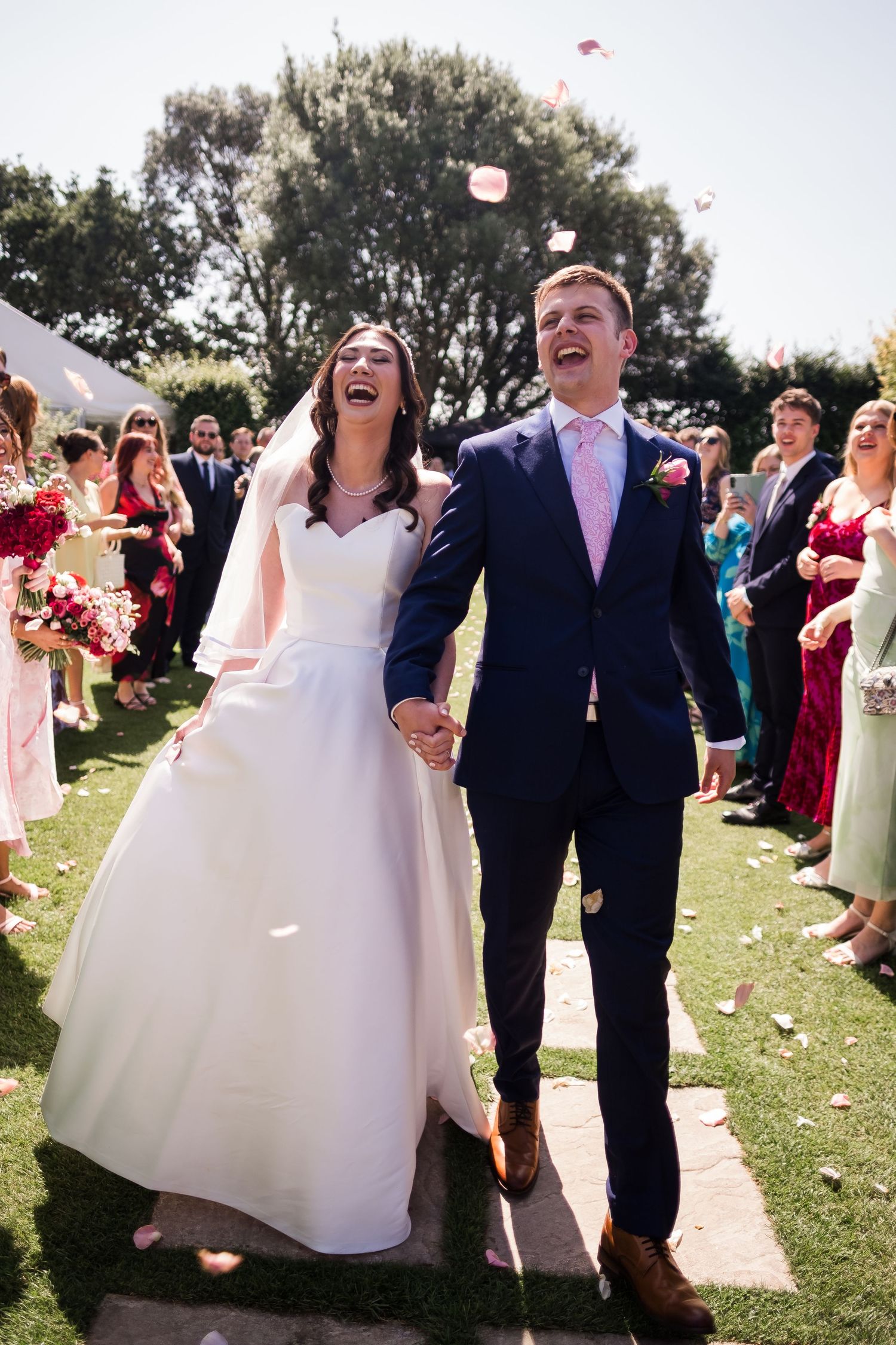 Wedding couple walks down aisle while guests toss flower petals in outdoor garden ceremony celebration.