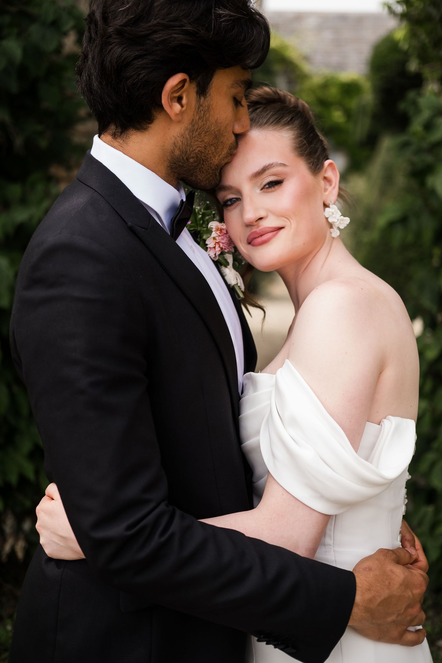 A groom in a black tuxedo kisses his bride in a white off-shoulder gown as they embrace outdoors.