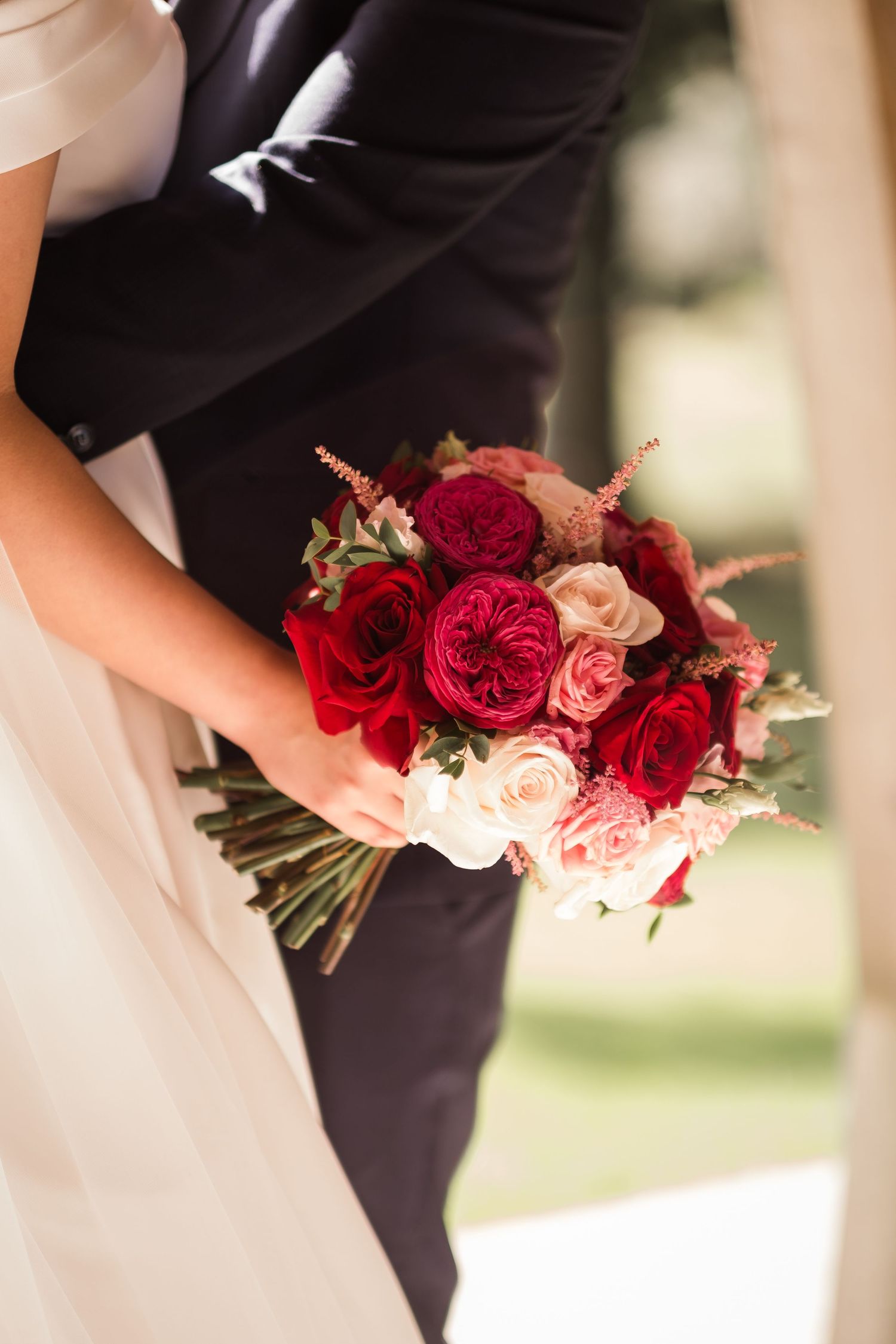 A romantic wedding bouquet featuring deep red and blush pink roses held against a black suit and white dress.
