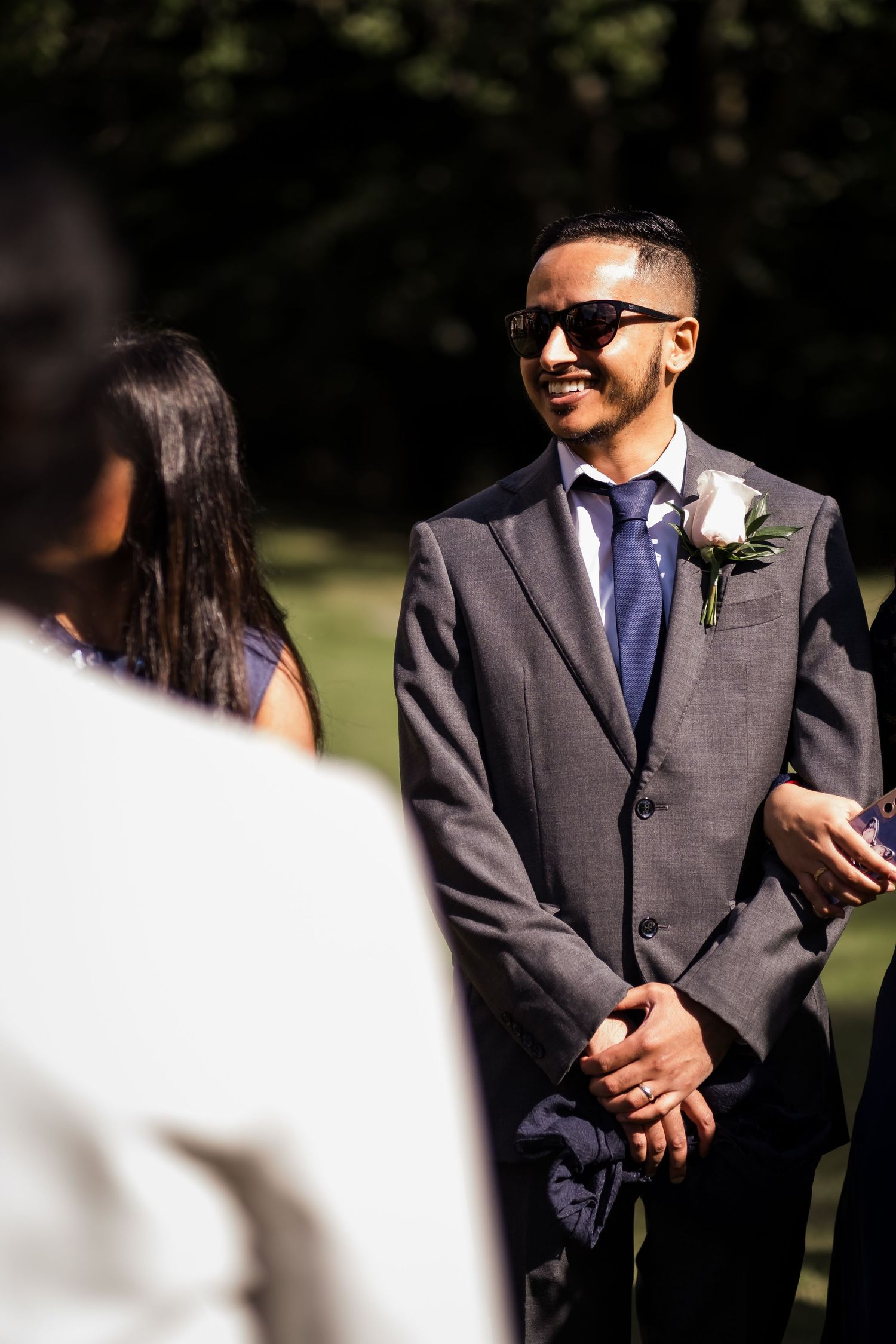 Groomsmen in dark suits with white boutonnieres stand together during an outdoor wedding ceremony.
