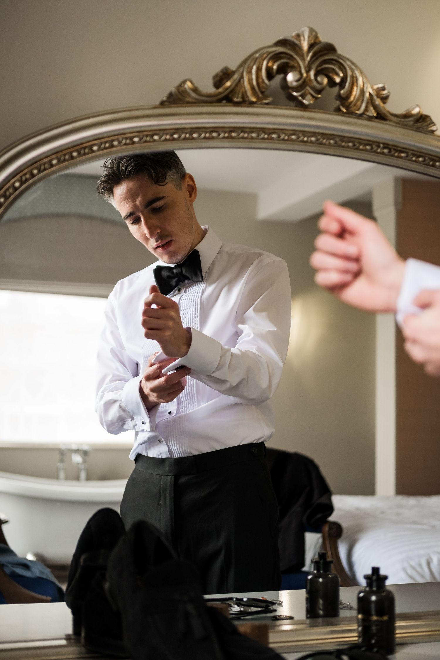 A person in formal attire sits at a mirrored vanity table in an elegant room with ornate molding details.