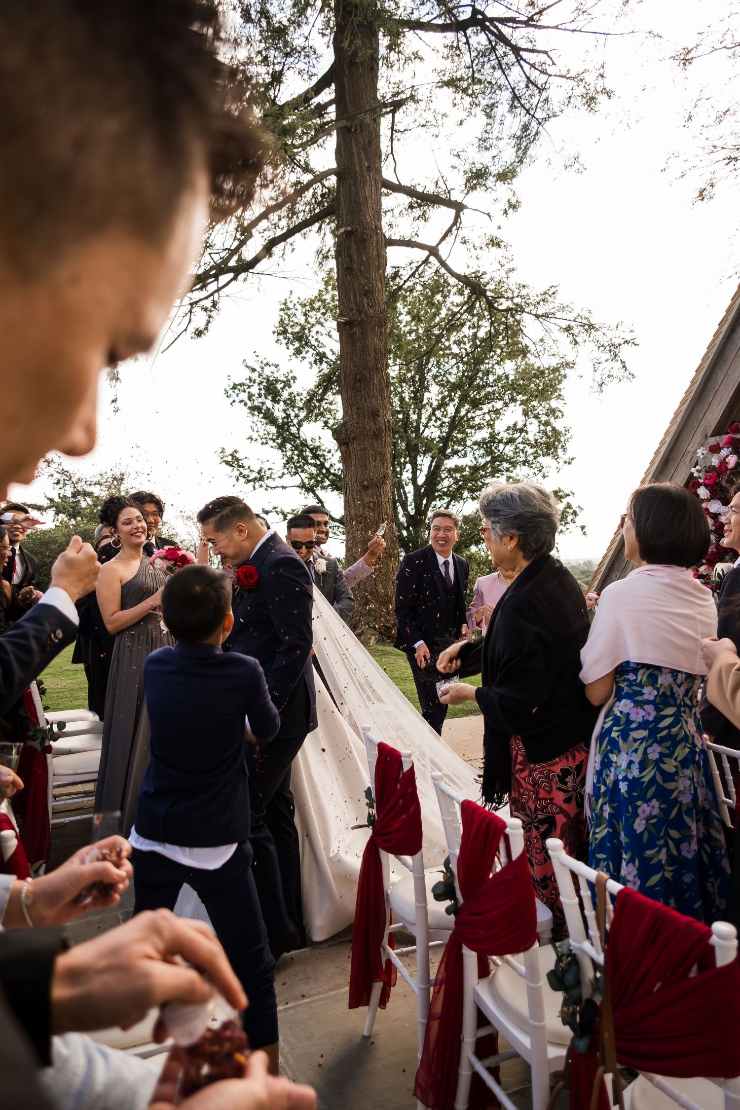 Wedding guests gather outdoors under trees during a reception ceremony in burgundy and navy attire.