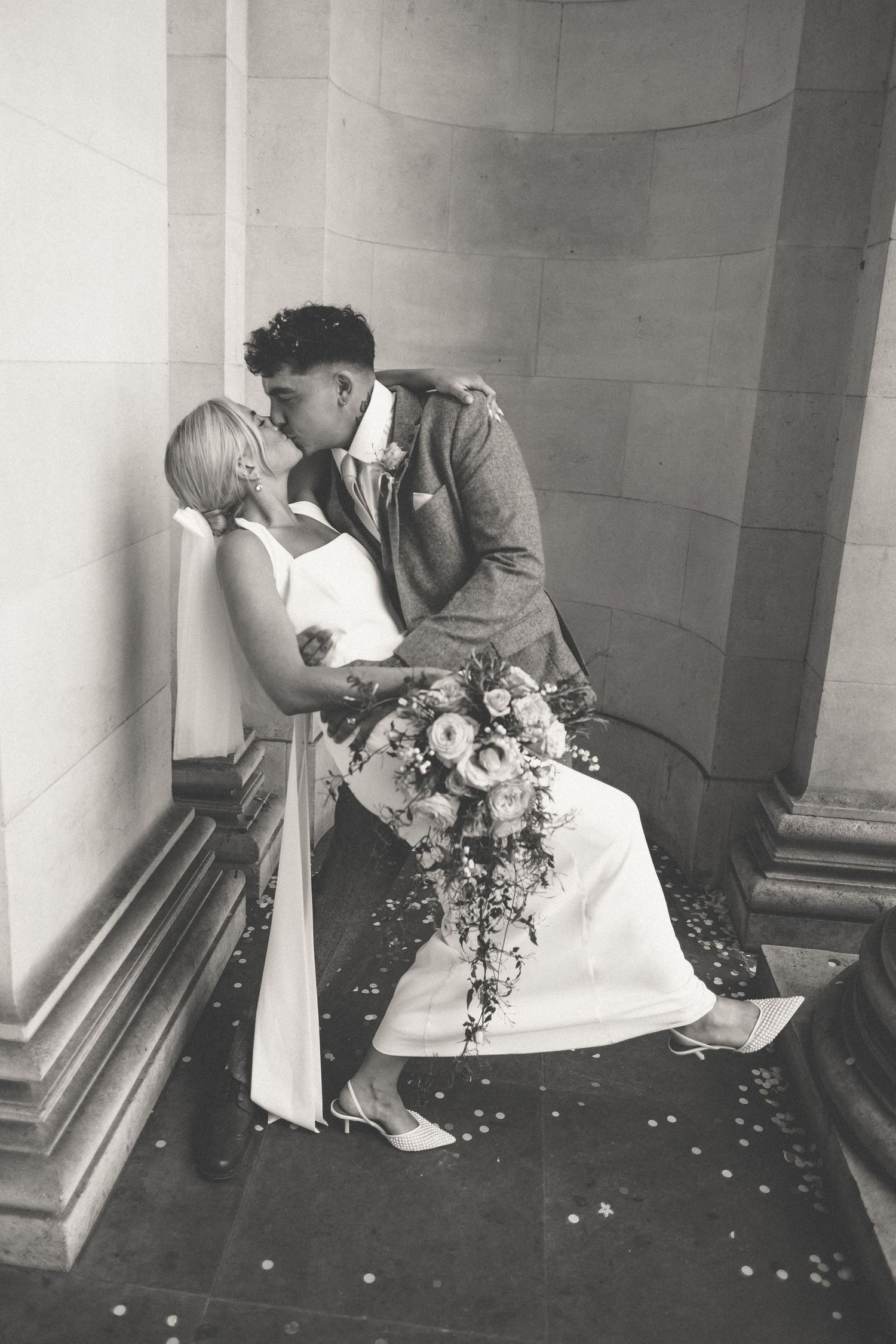 Black and white photo of a bride and groom sharing a romantic kiss on stone steps, bride holding a cascading floral bouquet.