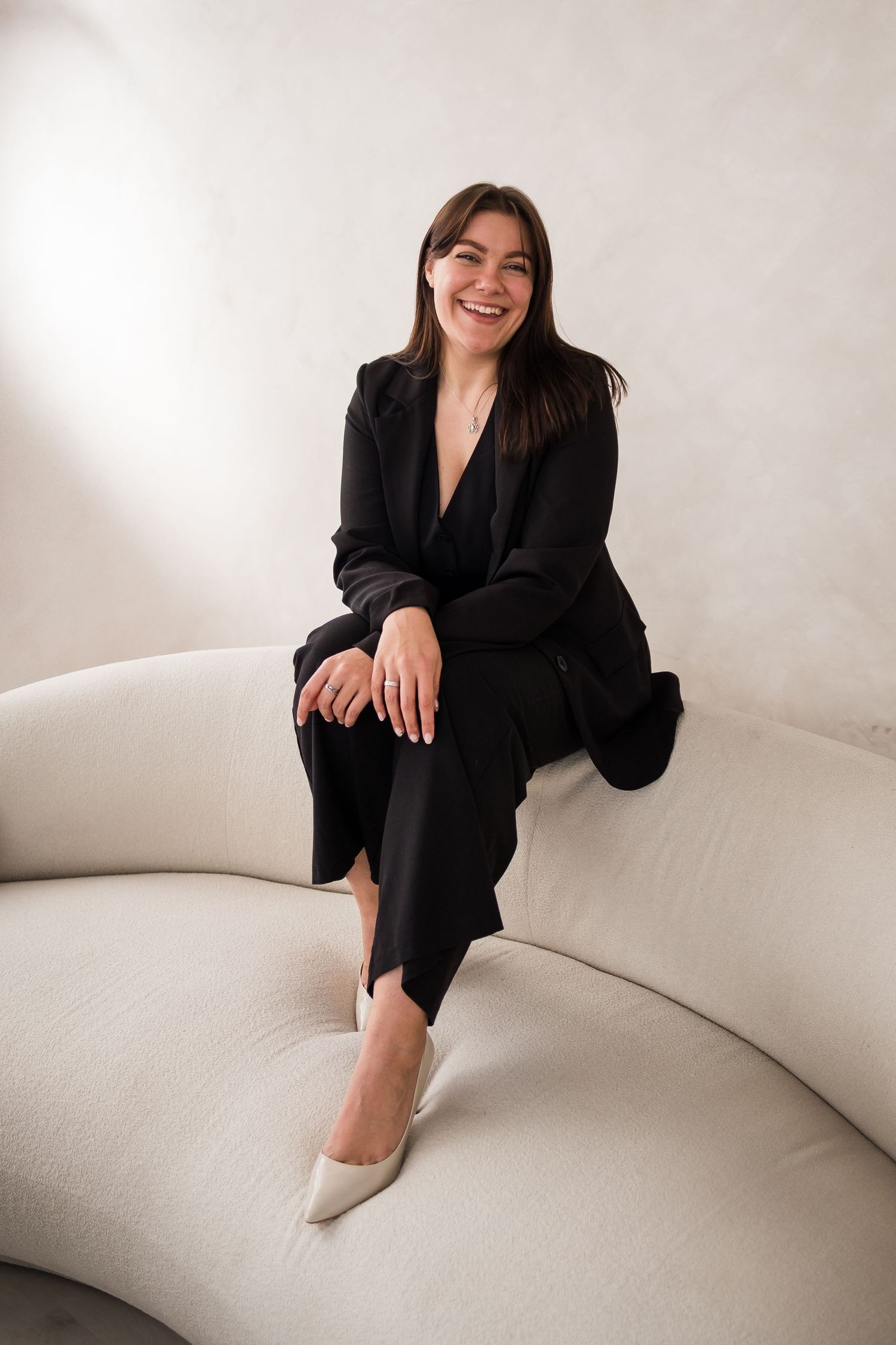 Professional lifestyle portrait on white curved sofa featuring black blazer and minimalist aesthetic.