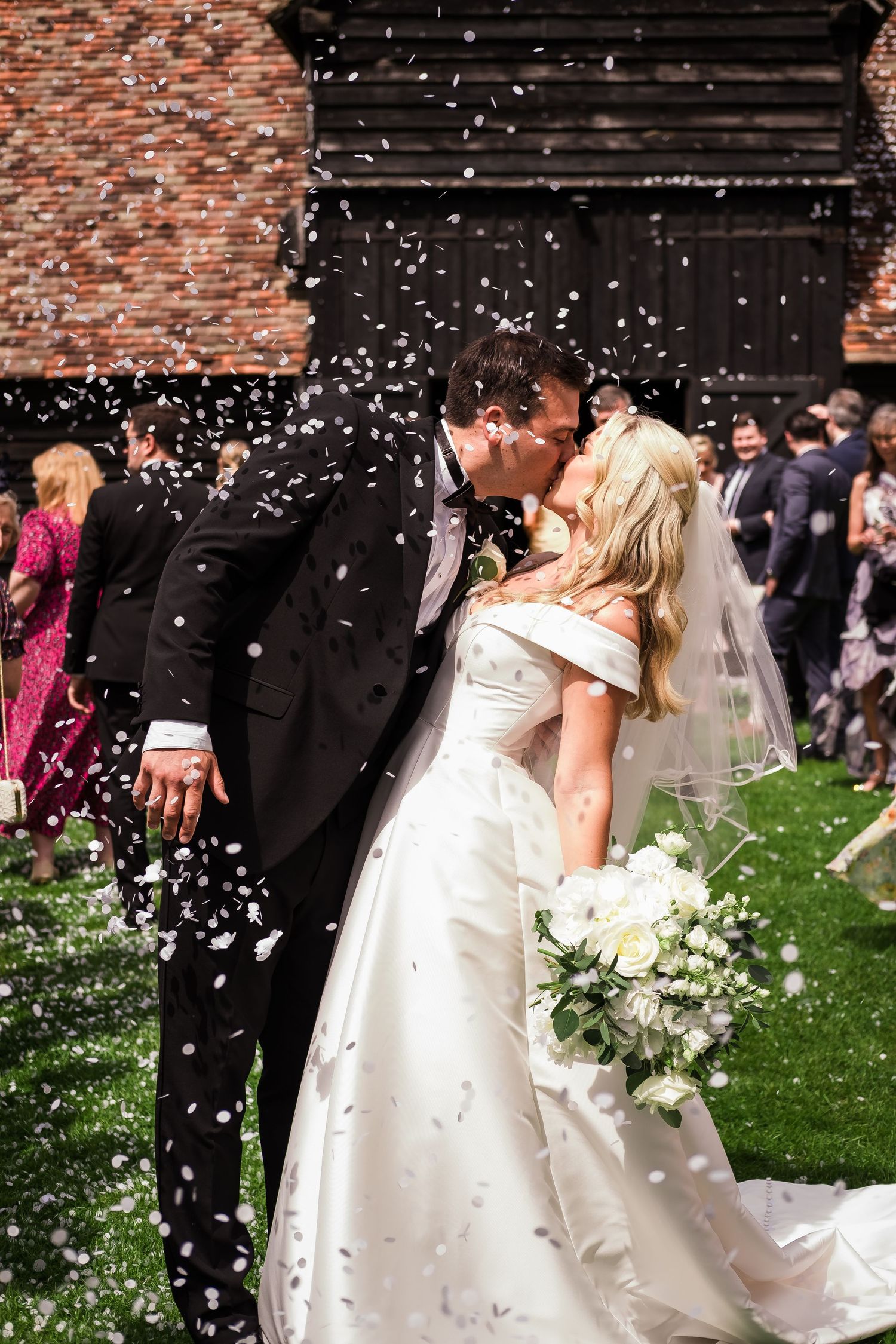 Bride and groom sharing a kiss surrounded by white confetti outside a rustic barn venue on their wedding day.