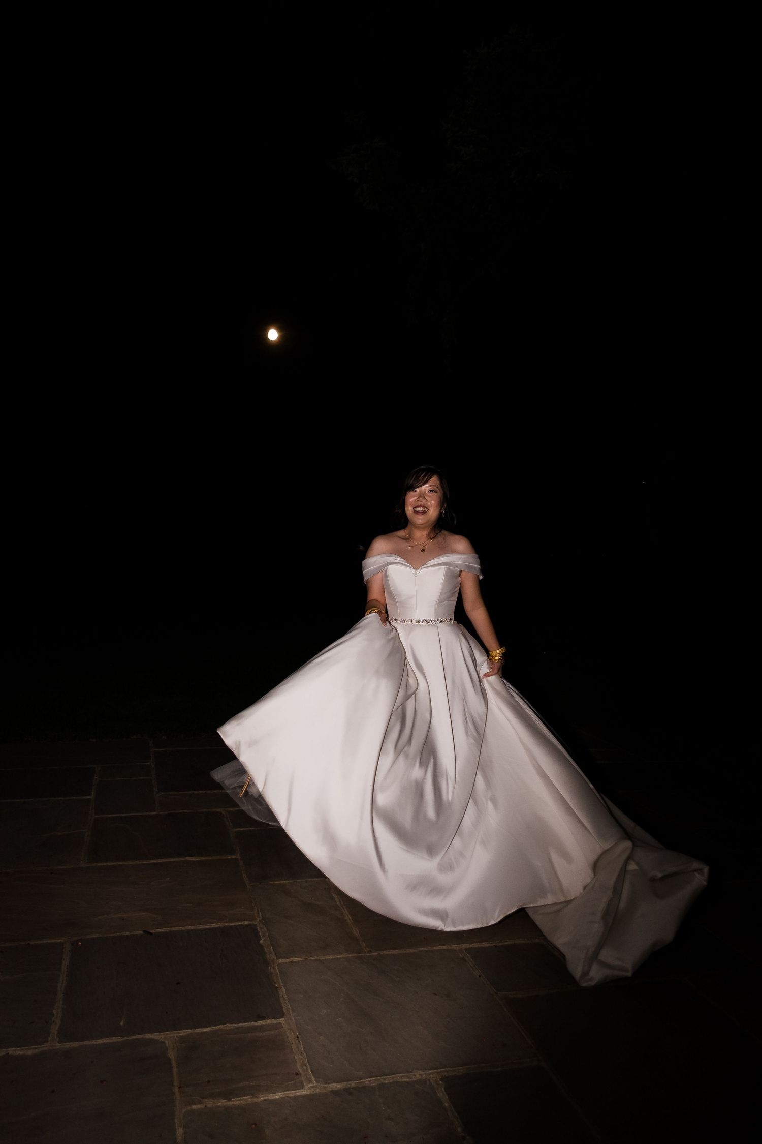 A person in an elegant white wedding gown twirls on dark stone pavement at night with a glowing moon in the background.