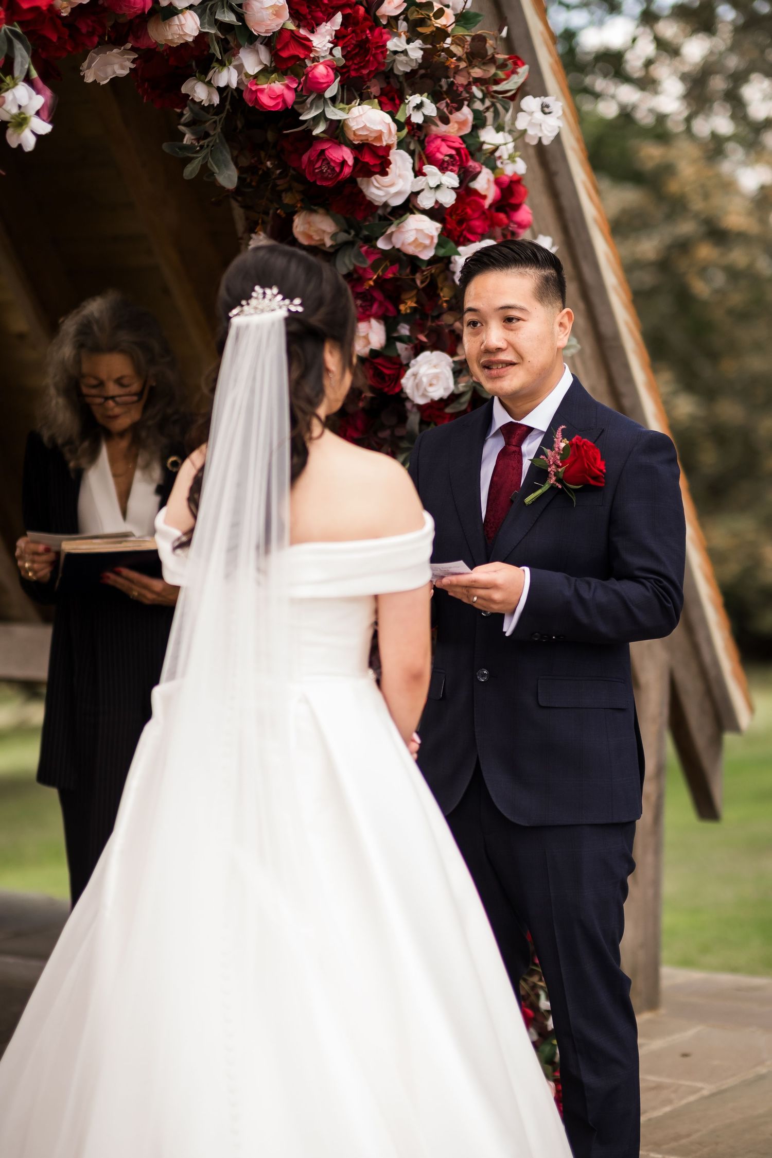 A couple exchanges vows under a wooden arch adorned with red and white roses during an outdoor wedding ceremony.