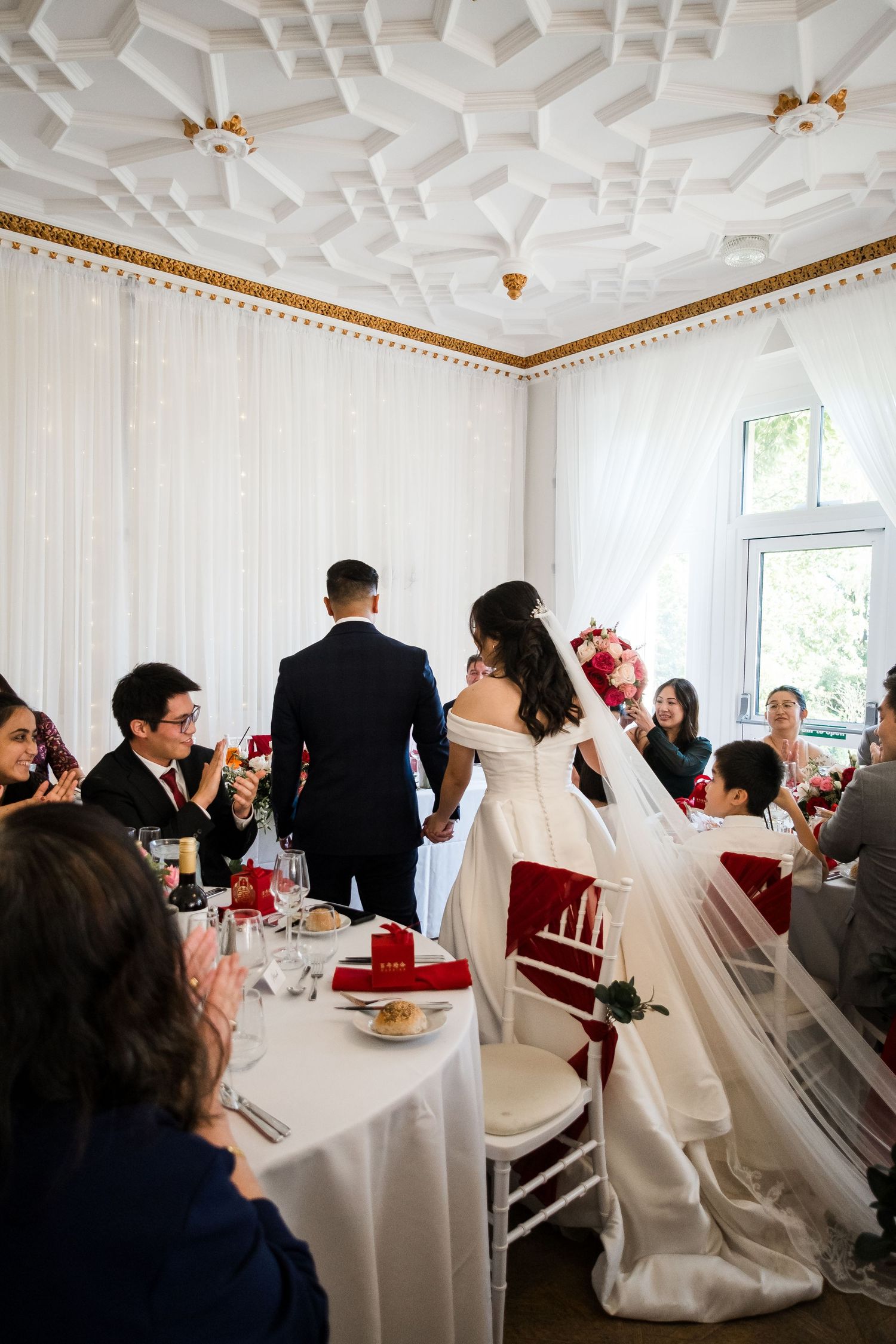 A wedding reception in a white room with ornate ceiling details and guests seated at elegant tables.