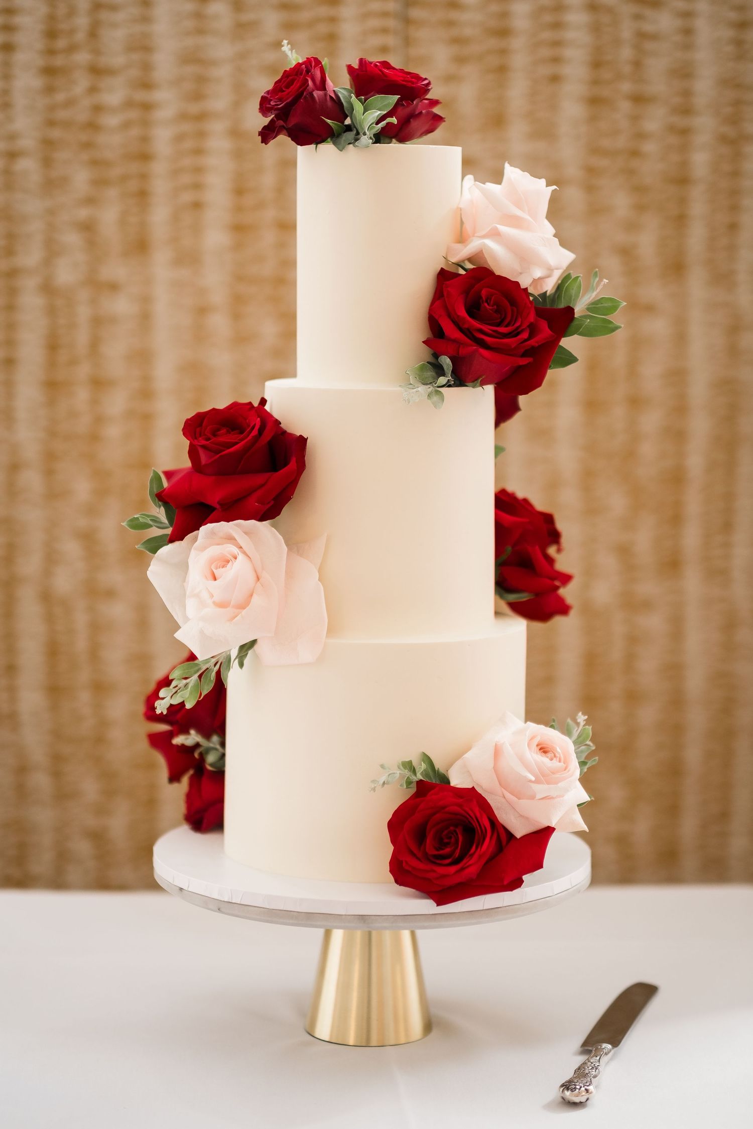 Elegant three-tier white wedding cake decorated with blush pink and deep red roses on a gold metallic cake stand.