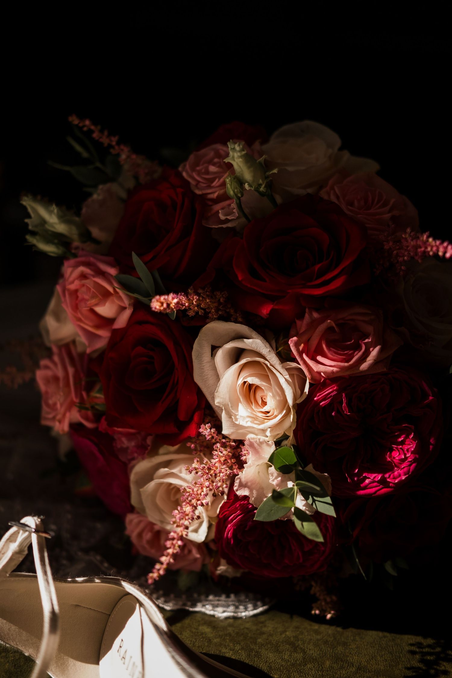 Romantic floral arrangement of dark red and blush pink roses with ribbon accents in moody lighting against black background.