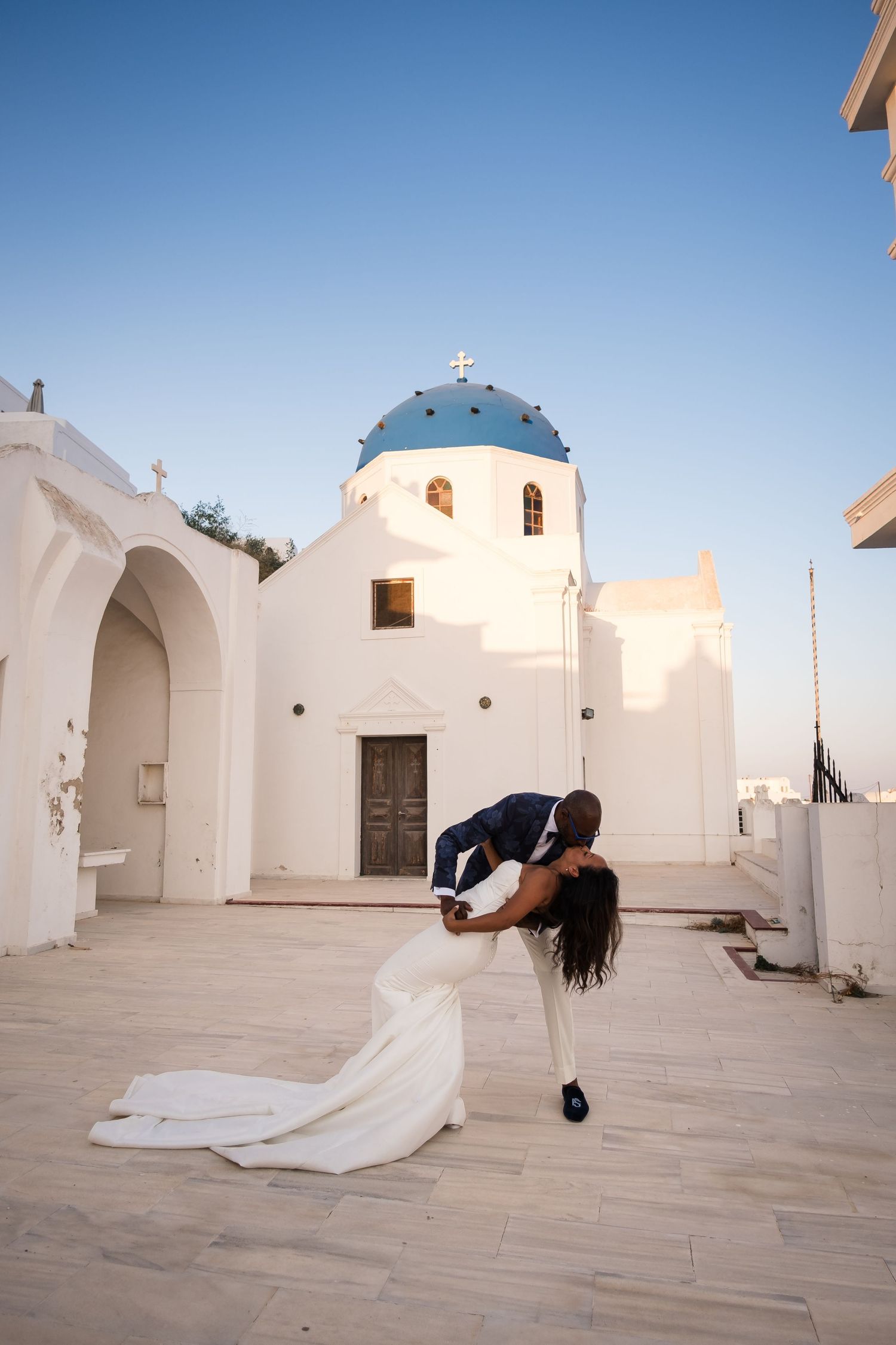 Couple sharing a romantic dip kiss in front of a white Greek Orthodox church with blue dome in Santorini.
