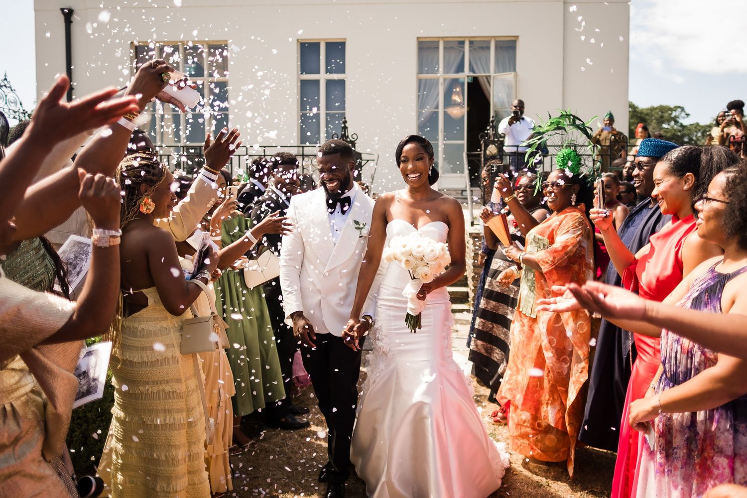 Joyful newlywed couple walks through confetti shower outside church, bride in white gown holding bouquet, groom in white tuxedo.
