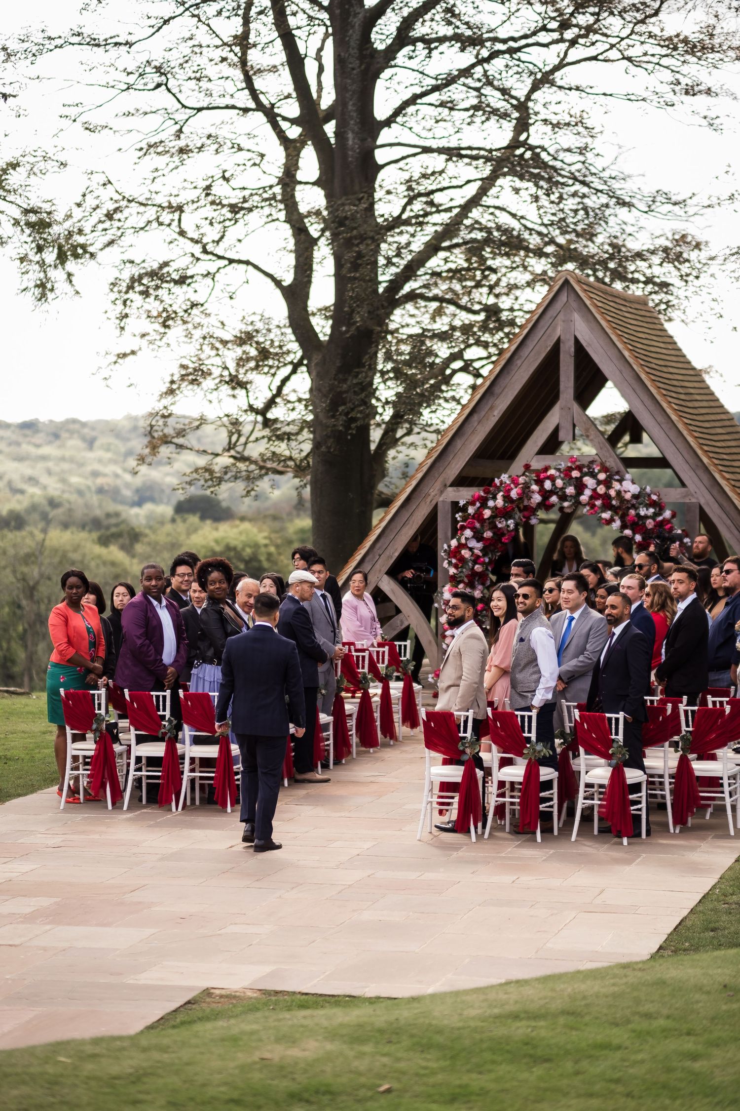 Guests seated in white chairs beneath wooden pavilions for an outdoor wedding ceremony decorated with burgundy accents.
