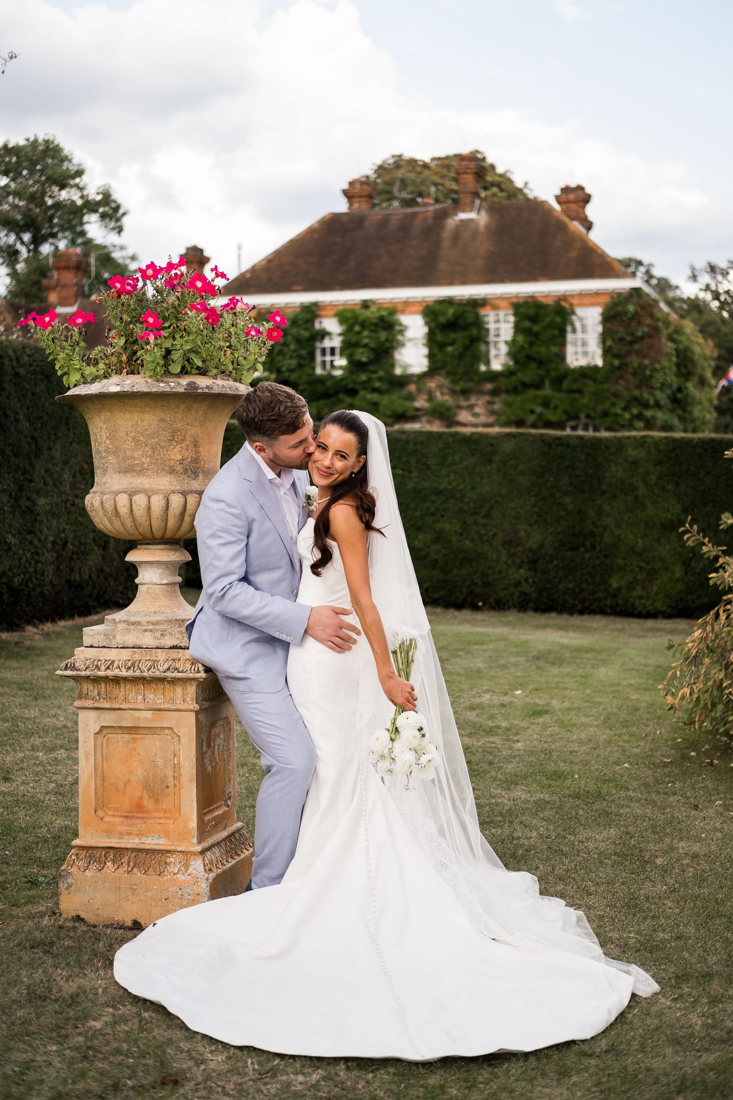 A couple in formal wedding attire embraces on a manicured lawn with stone urns and hedges in the background.