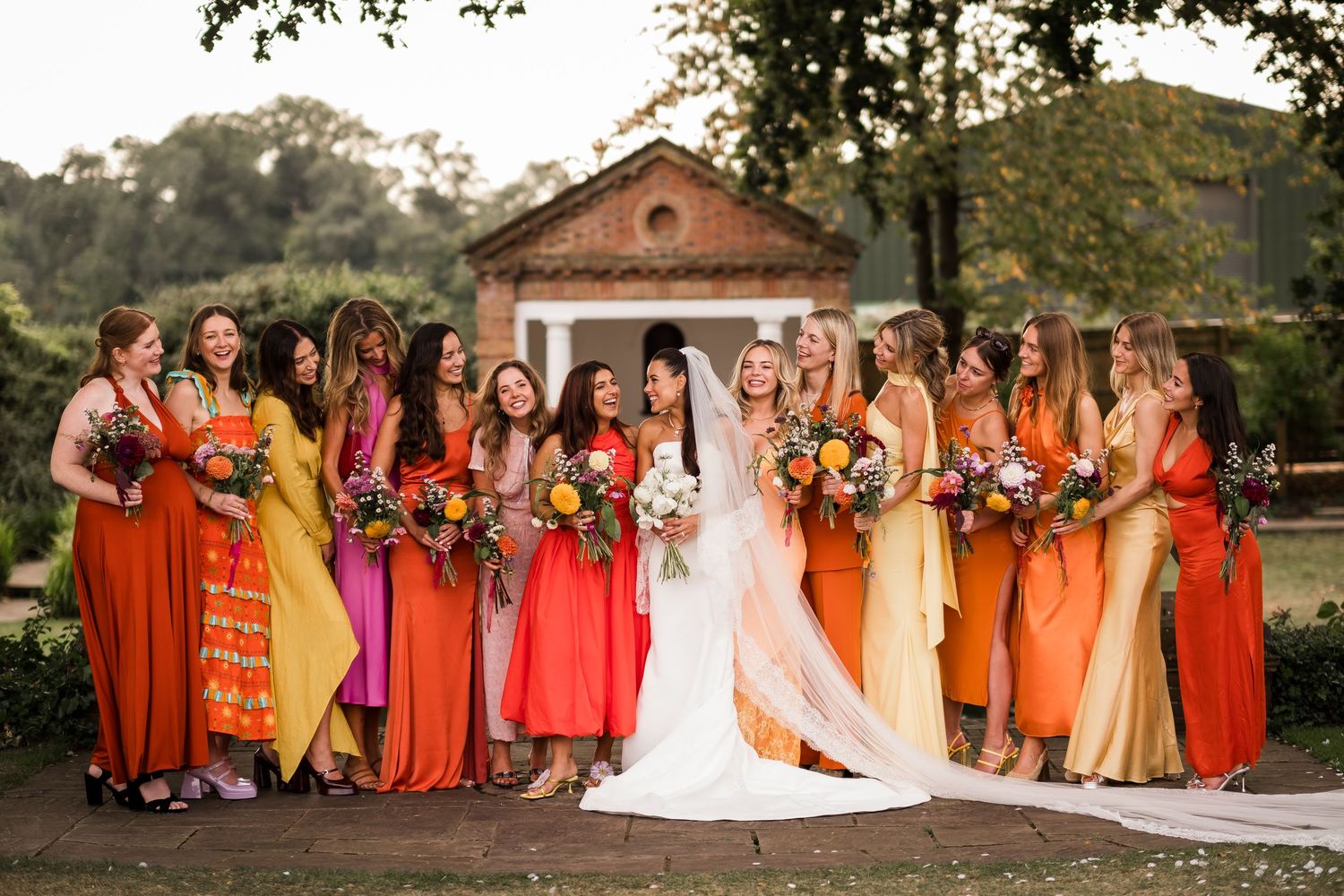 Bride in white gown surrounded by bridesmaids in mismatched orange, red, and yellow dresses holding colorful bouquets outdoors.
