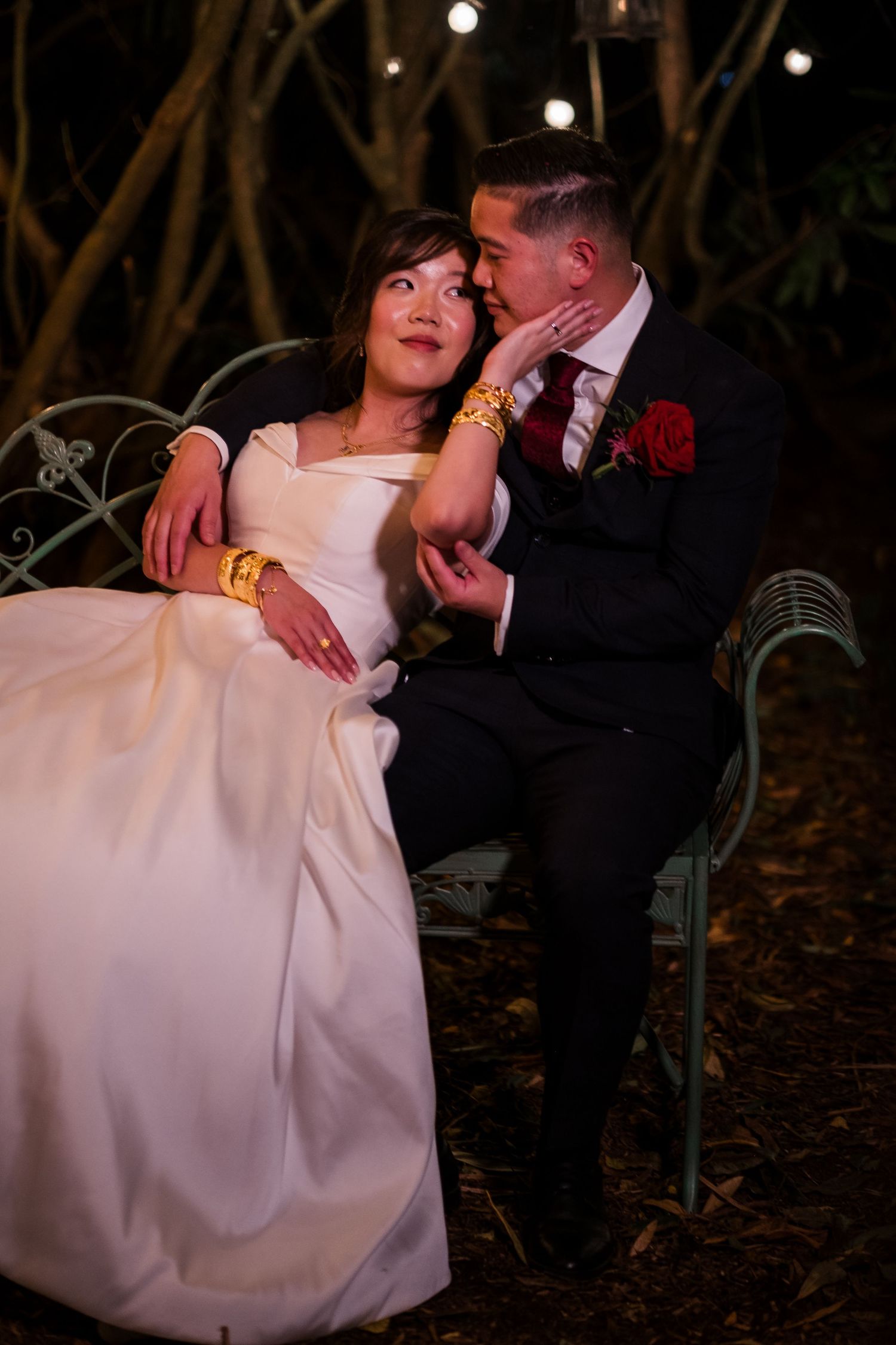 A romantic wedding couple shares an intimate moment on a garden bench under twinkling lights at night.